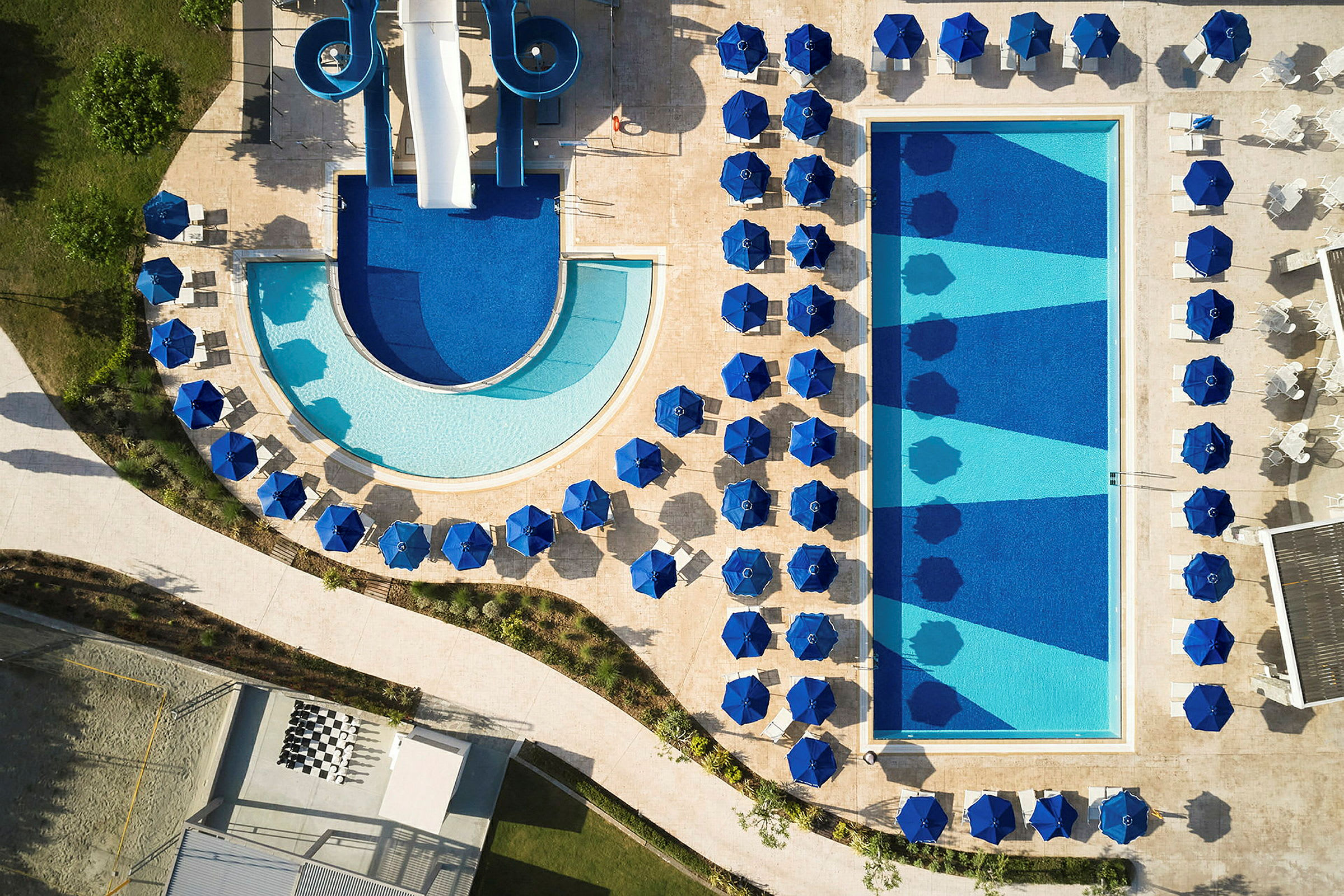 Flygvy över en swimmingpool samt blå parasoller och solstolar runtom. (AI-genererad text)