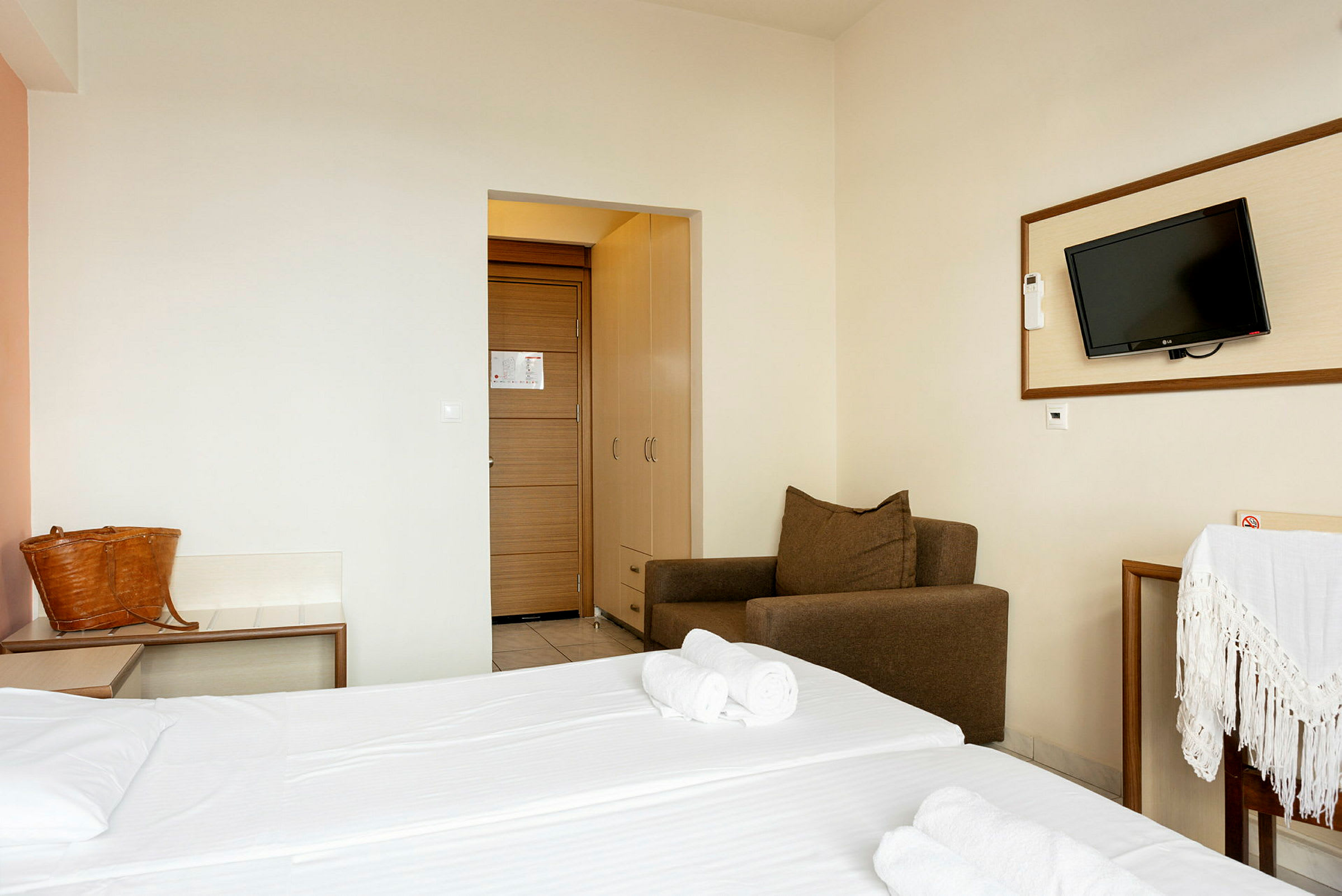 Hotellrum med dubbelsäng, brun fåtölj, upphängd TV och dörr med garderob. (AI-genererad text)