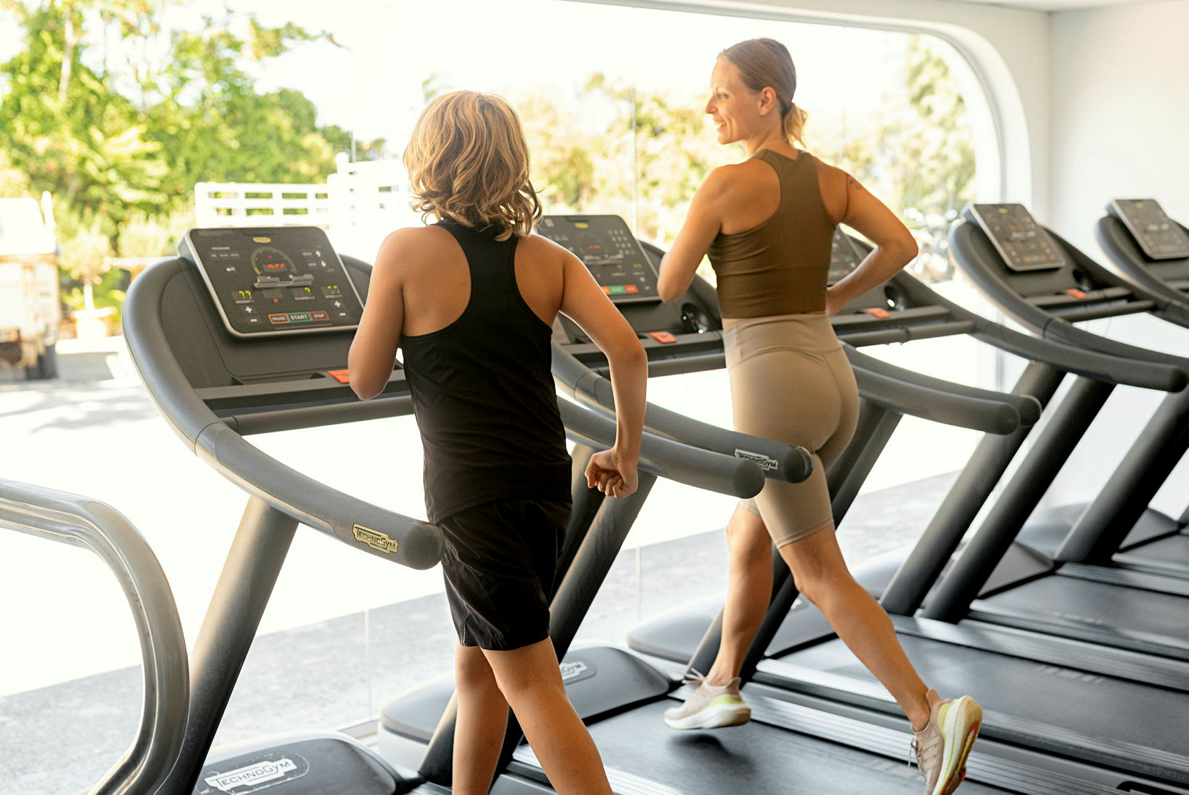 Två personer som tränar på löpband i ett gym med stora fönster och solljus. (AI-genererad text)