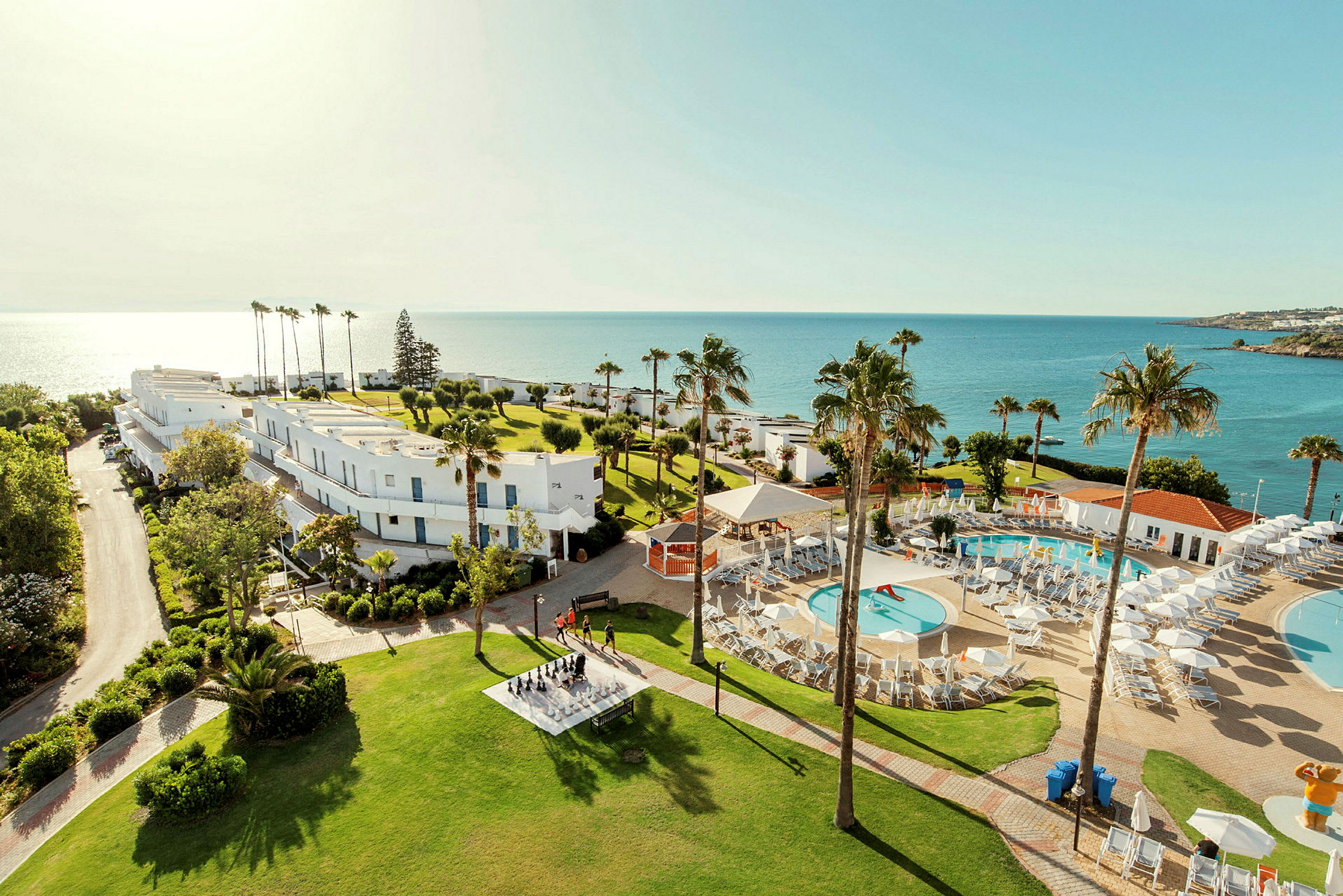 En resort vid havet med poolområde, gröna gräsmattor och vita byggnader. (AI-genererad text)