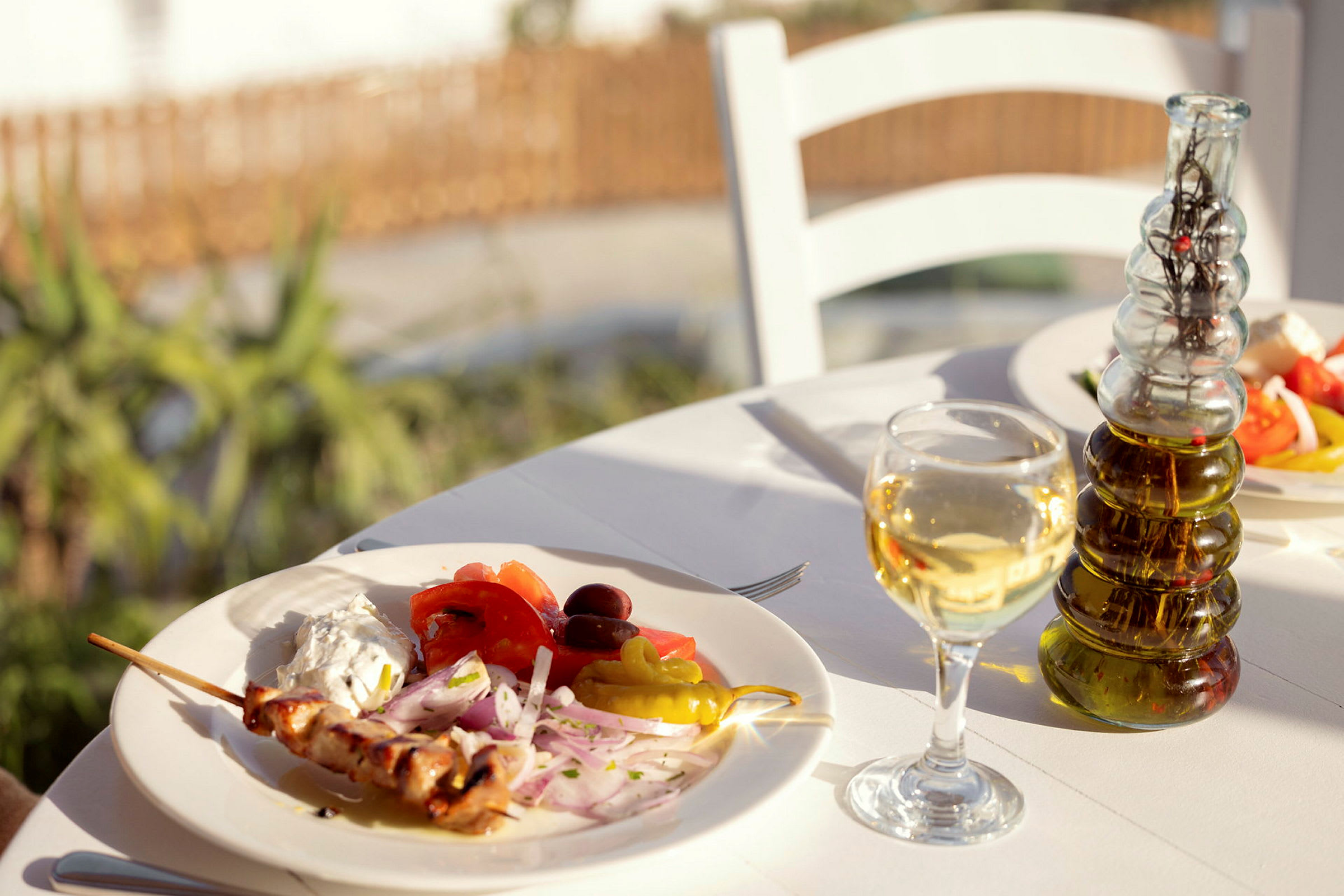 En tallrik med grillspett, grönsaker och sås på ett bord utomhus, med ett glas vitt vin och en flaska olivolja. (AI-genererad text)