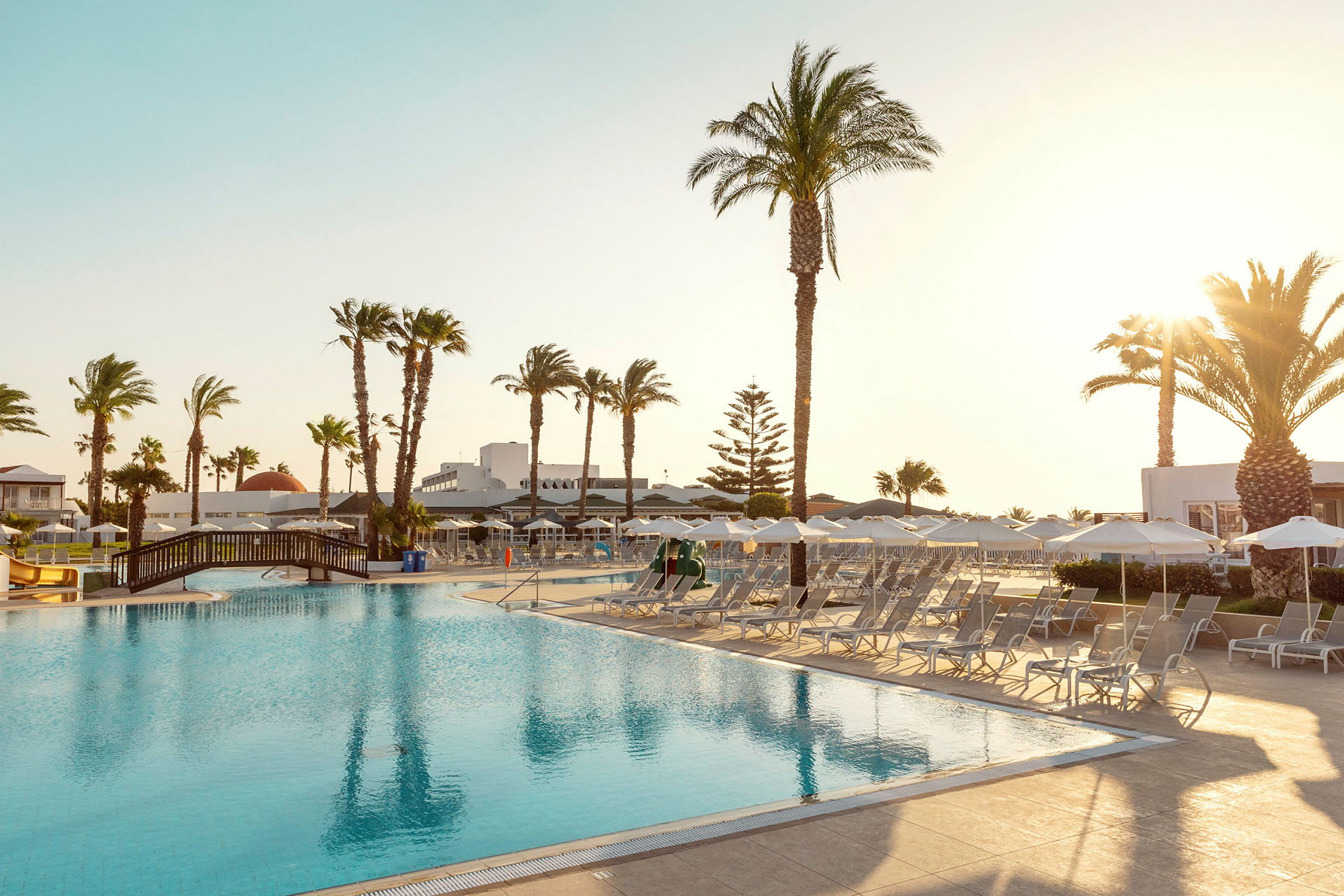 En hotellpool omgiven av solstolar och parasoller, med palmer och byggnader i bakgrunden. (AI-genererad text)