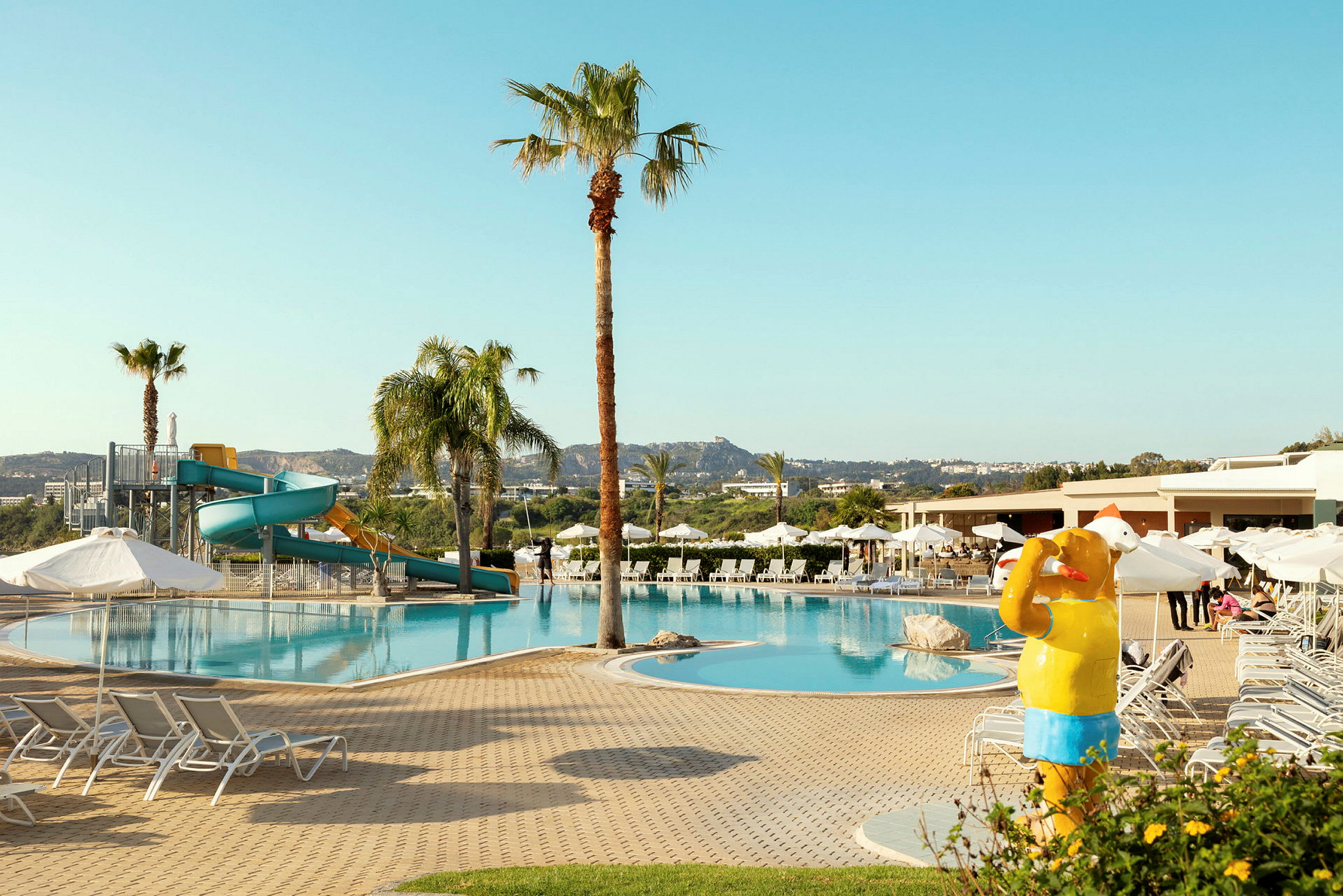 En pool med solstolar, en lekplats med vattenrutschbana och en gul skulptur föreställande en badande person. (AI-genererad text)