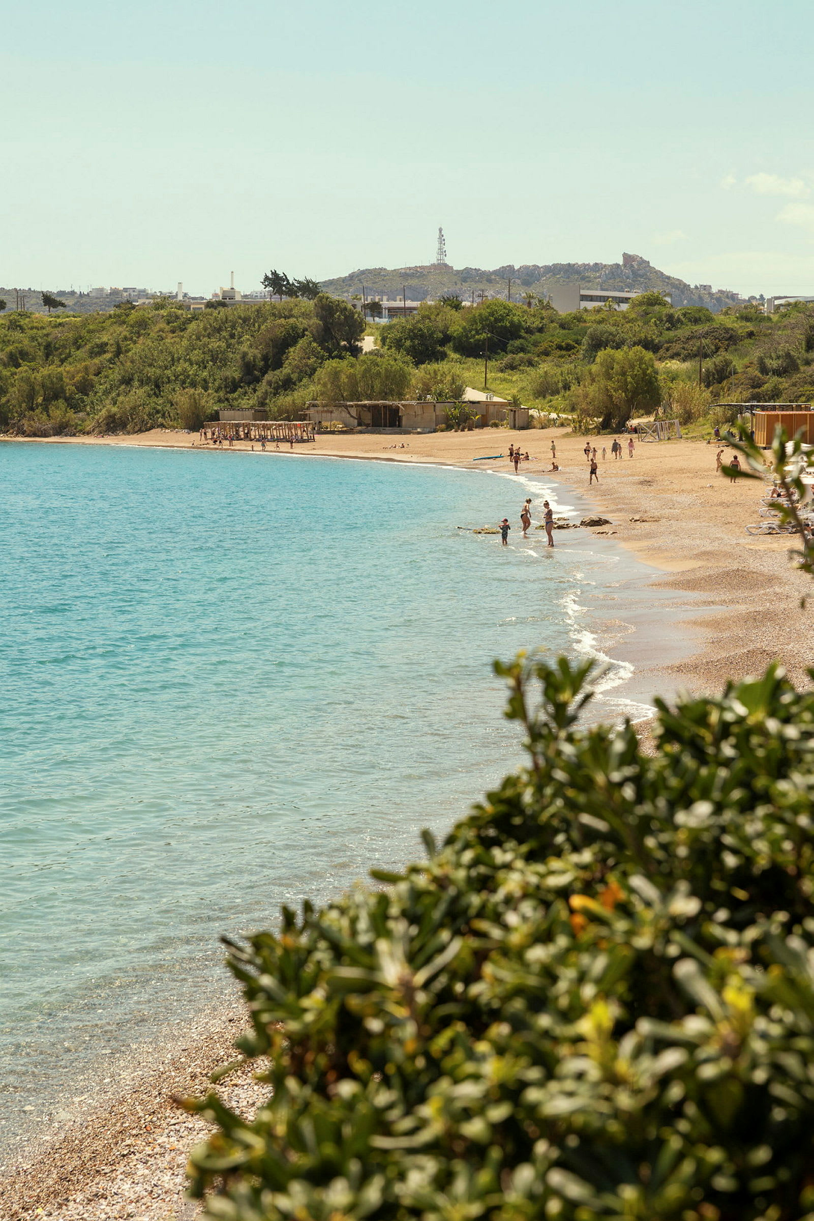 Vy över en strand med turkost vatten och badande människor, omgiven av grönska. (AI-genererad text)