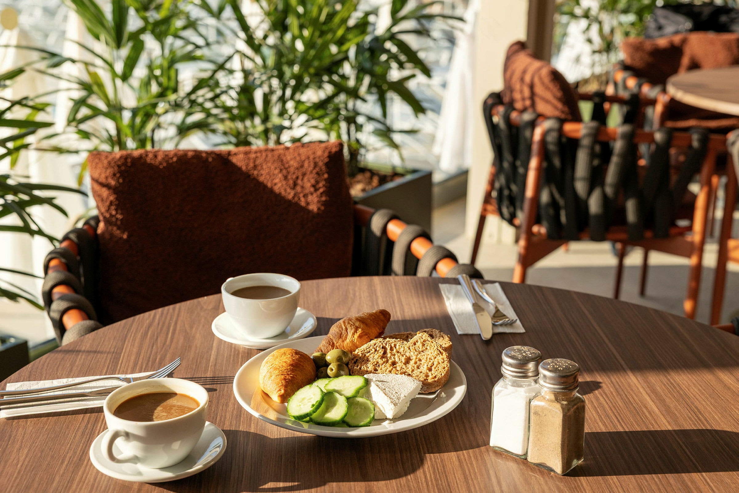 En frukostuppsättning med kaffe och en tallrik med bröd, ost, gurkskivor och en croissant på ett bord. (AI-genererad text)
