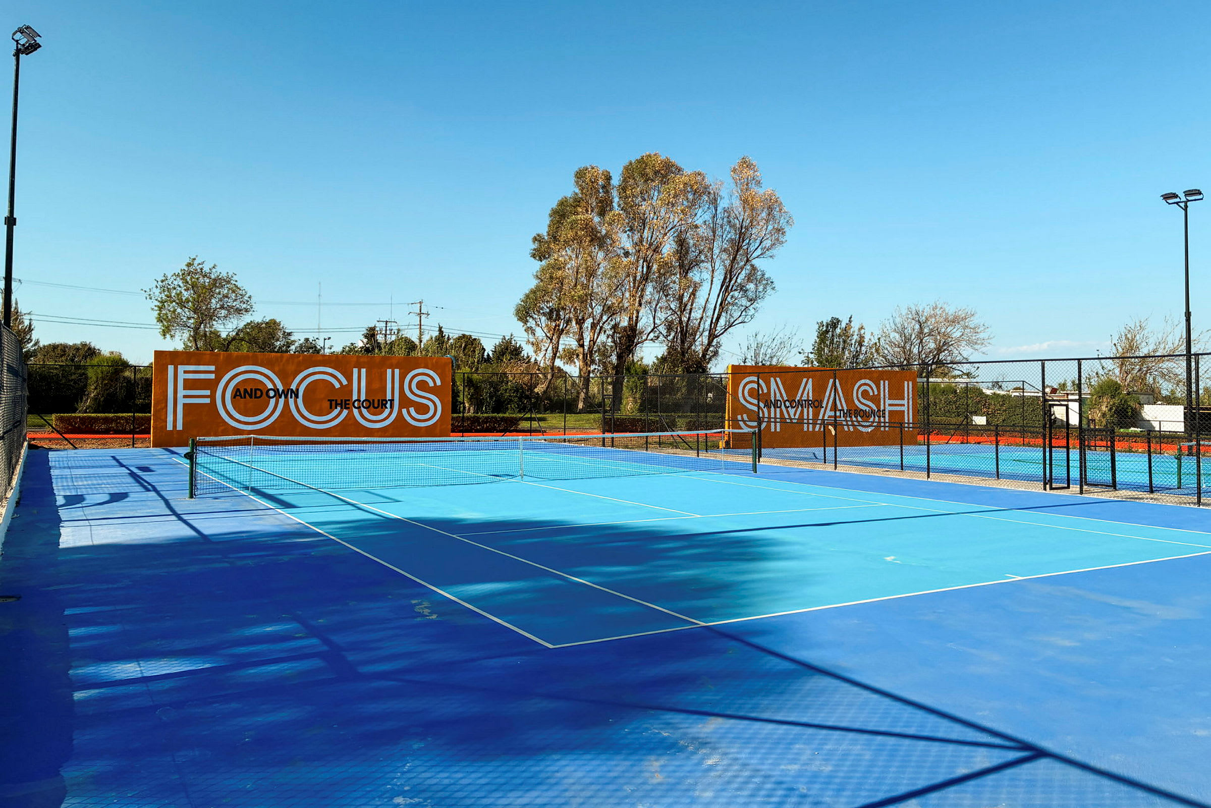 Blå tennisbana utomhus med träd i bakgrunden och skyltar som visar texten "Focus" och "Smash". (AI-genererad text)