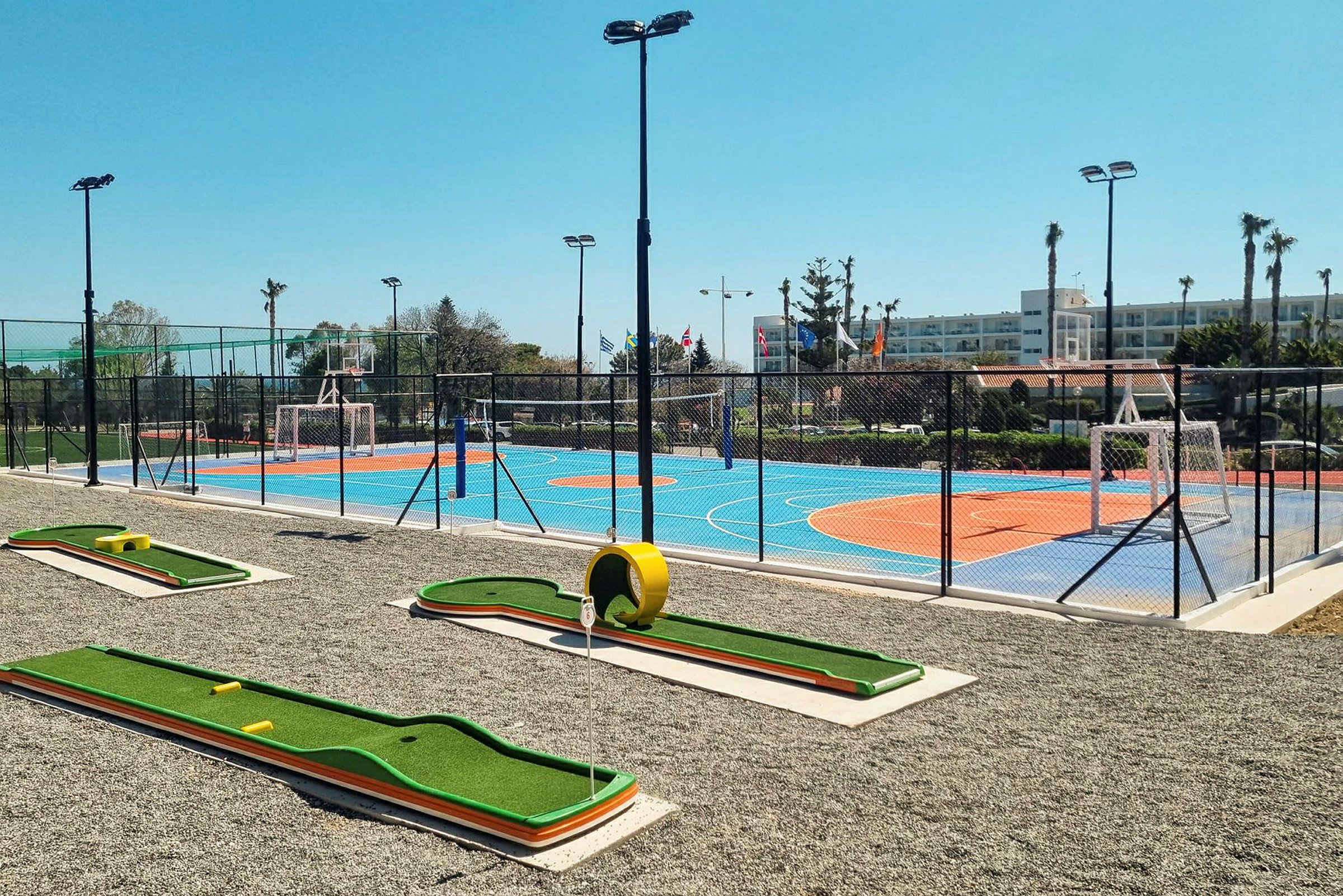 En bild av en utomhusmiljö med en minigolfbana i förgrunden och en färgglad sportplan med orange och blå yta omgiven av stängsel i bakgrunden. (AI-genererad text)