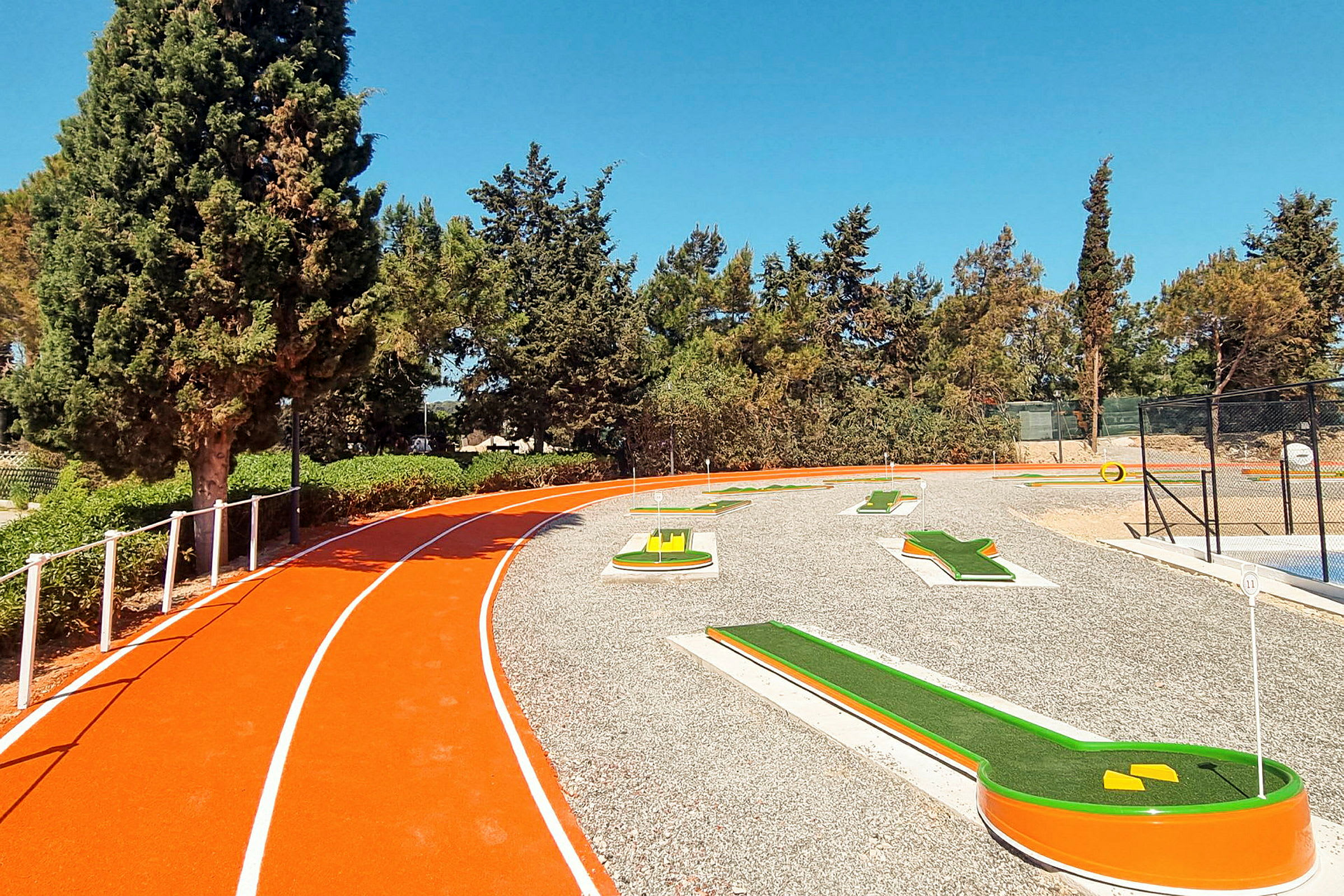 En bild av en minigolfbana bredvid en orange löparbana. Bilden är tagen utomhus med grön vegetation och klarblå himmel i bakgrunden. (AI-genererad text)