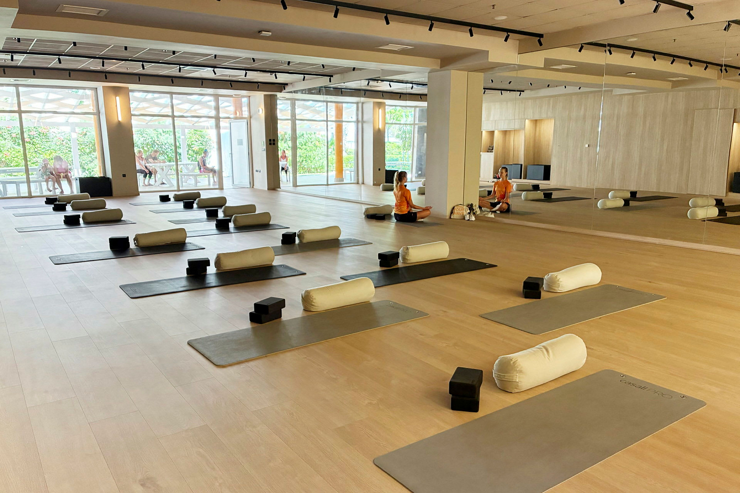 Ett träningsrum med yogamattor och tillbehör som kuddar och block uppställda. Två personer samtalar vid spegeln i bakgrunden. (AI-genererad text)