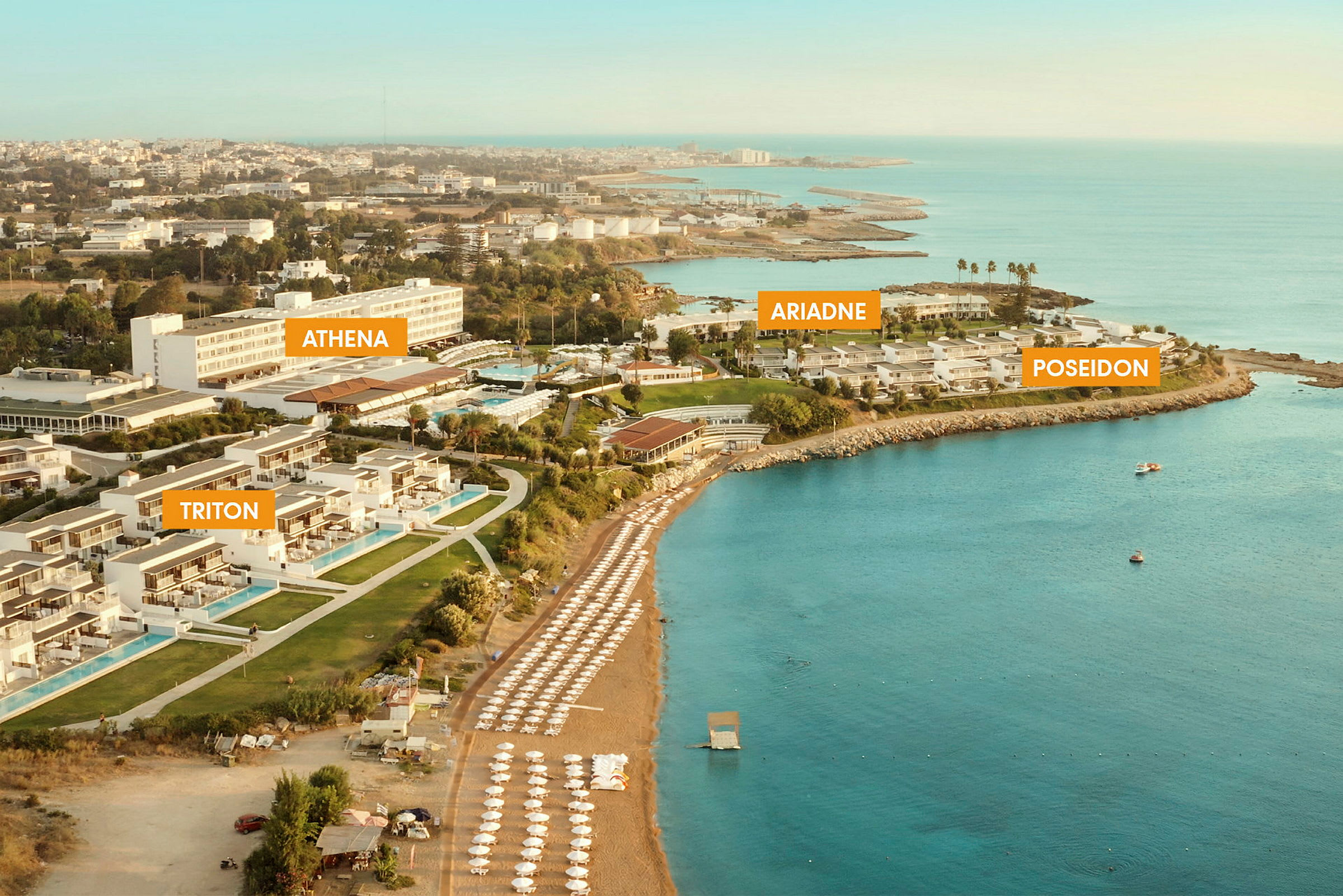 Flygfoto över ett hotellområde nära kusten med flera byggnader och en lång sandstrand. (AI-genererad text)