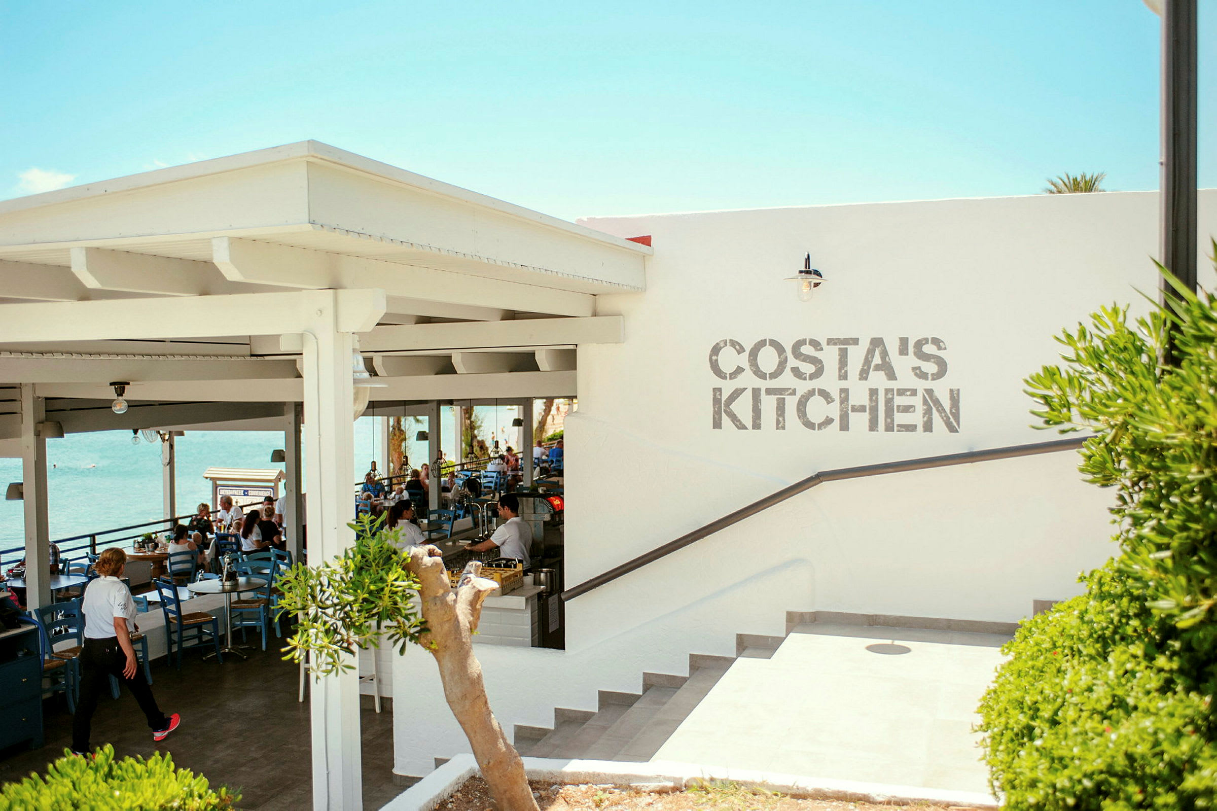 En utomhusrestaurang med havsutsikt och texten 'Costa's Kitchen' på väggen. (AI-genererad text)