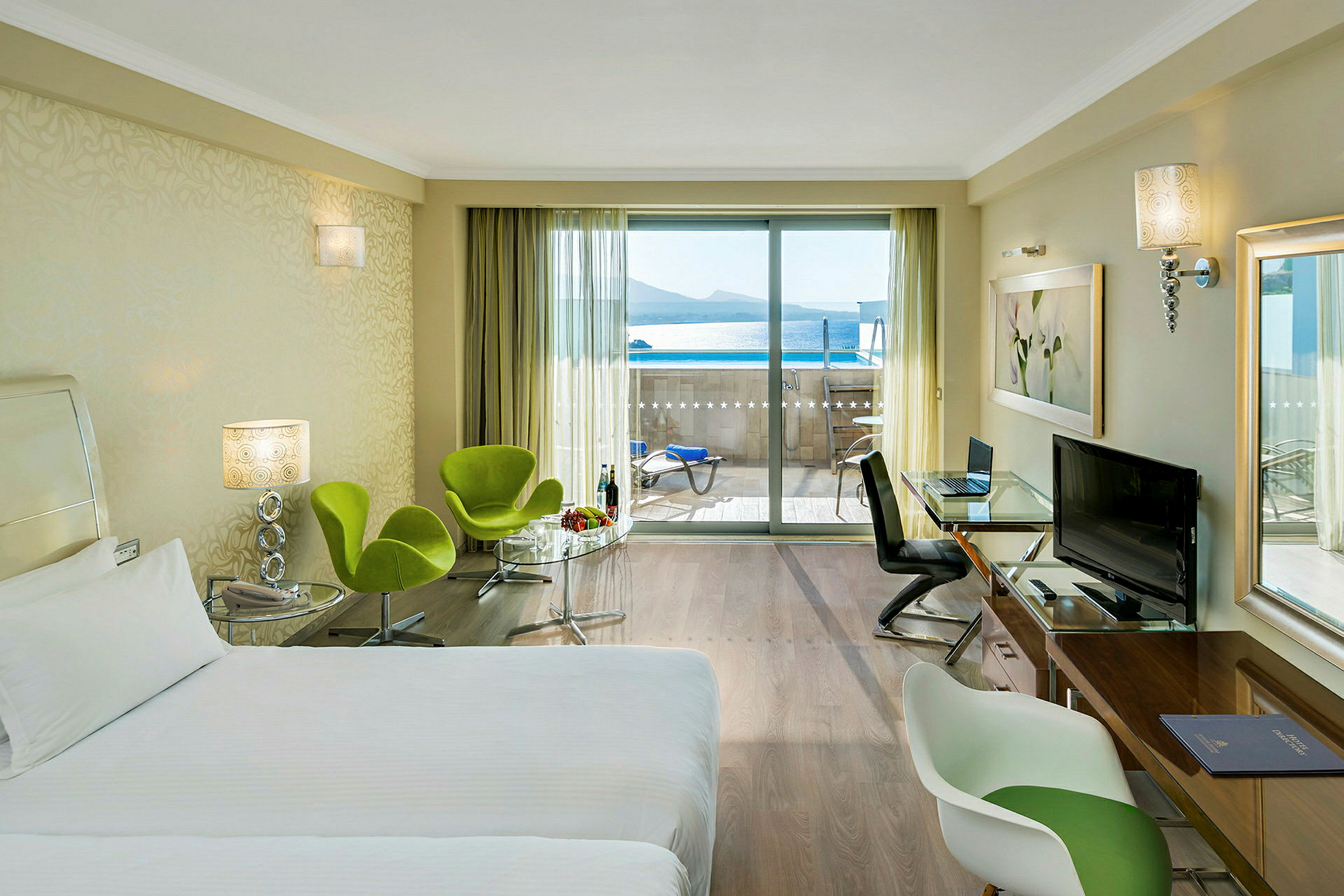 Ett hotellrum med en säng, sittmöbler och ett bord, samt utsikt mot en balkong med havsutsikt. (AI-genererad text)