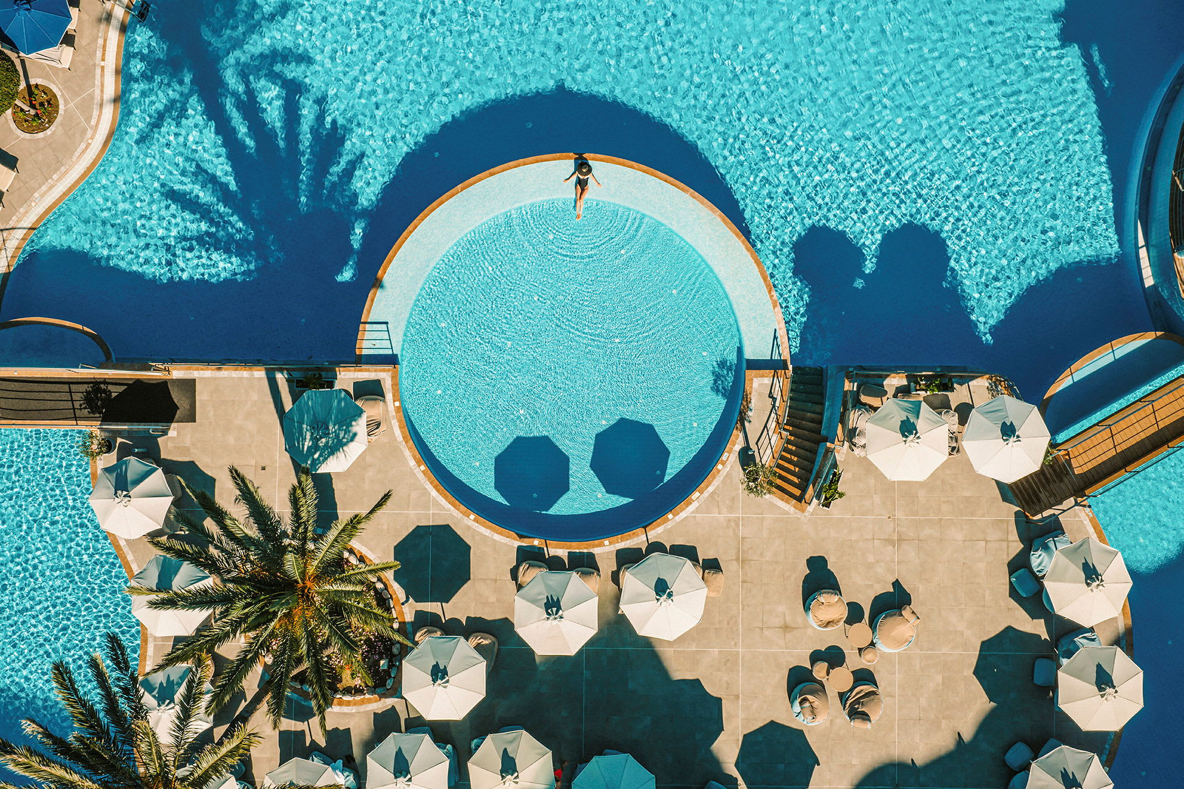 Luftfoto av en pool och solområdet, med parasoller, palmer och badande. (AI-genererad text)