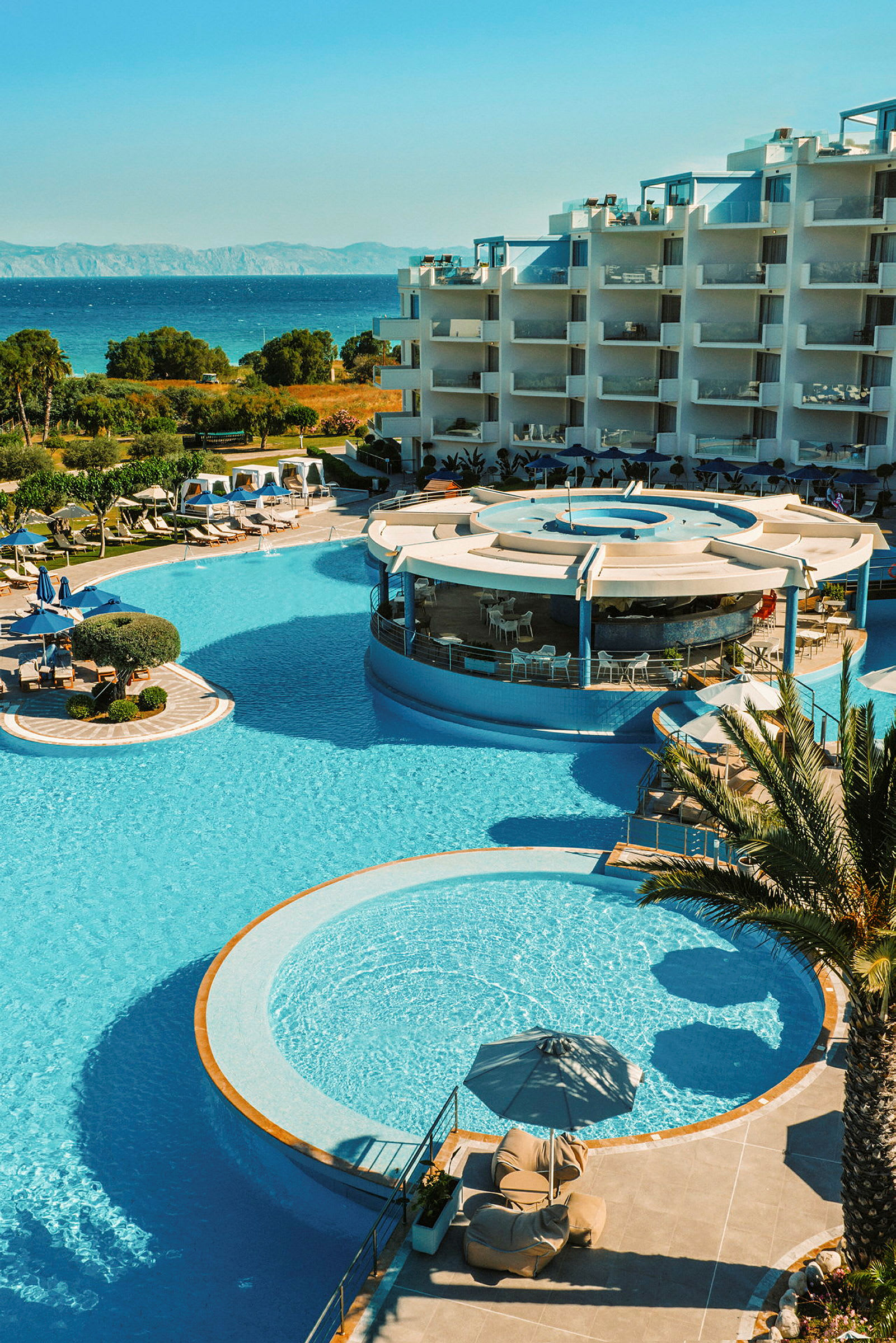 Bilden visar en semesterresort med en stor turkos pool, solstolar och parasoller, och en byggnad i modern stil. I bakgrunden syns havet och en bergig horisont. (AI-genererad text)