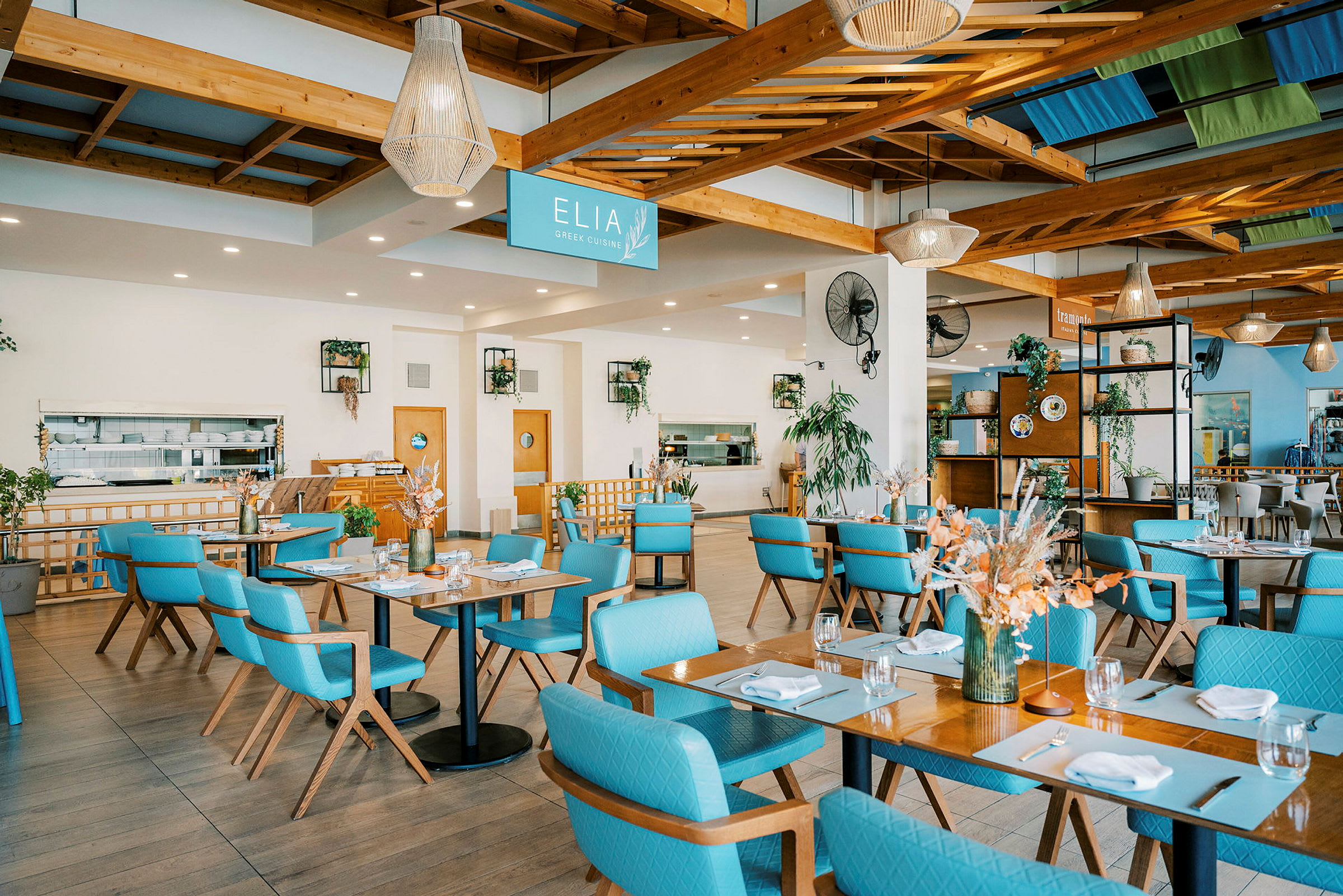 En ljus och rymlig restaurangmiljö med bord och blåa stolar. Dekorationer och grönska syns i inredningen. (AI-genererad text)