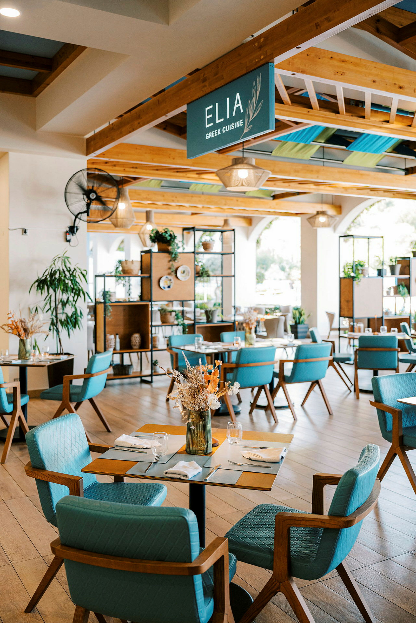 En ljus och modern restaurang med trädetaljer och blå stolar. Dekorerad med växter och torkade blomsterarrangemang. (AI-genererad text)