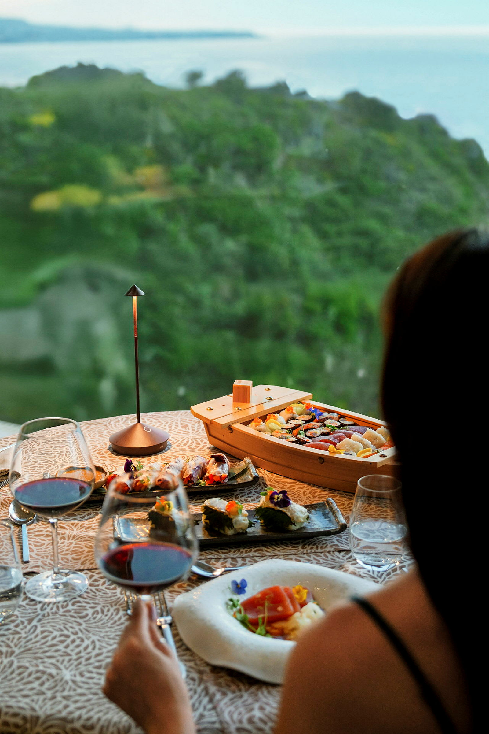En vackert dukad måltid med sushi och vin står på ett bord med utsikt över en grön landskap och havet. (AI-genererad text)
