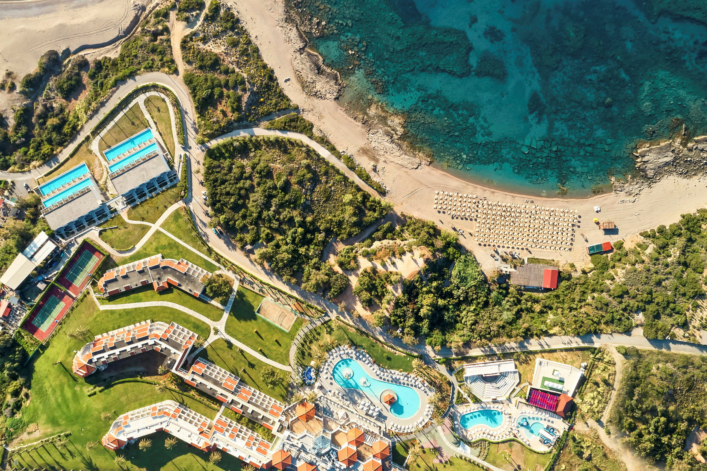 Flygfoto av ett hotellområde med pooler, byggnader, strand och hav. (AI-genererad text)