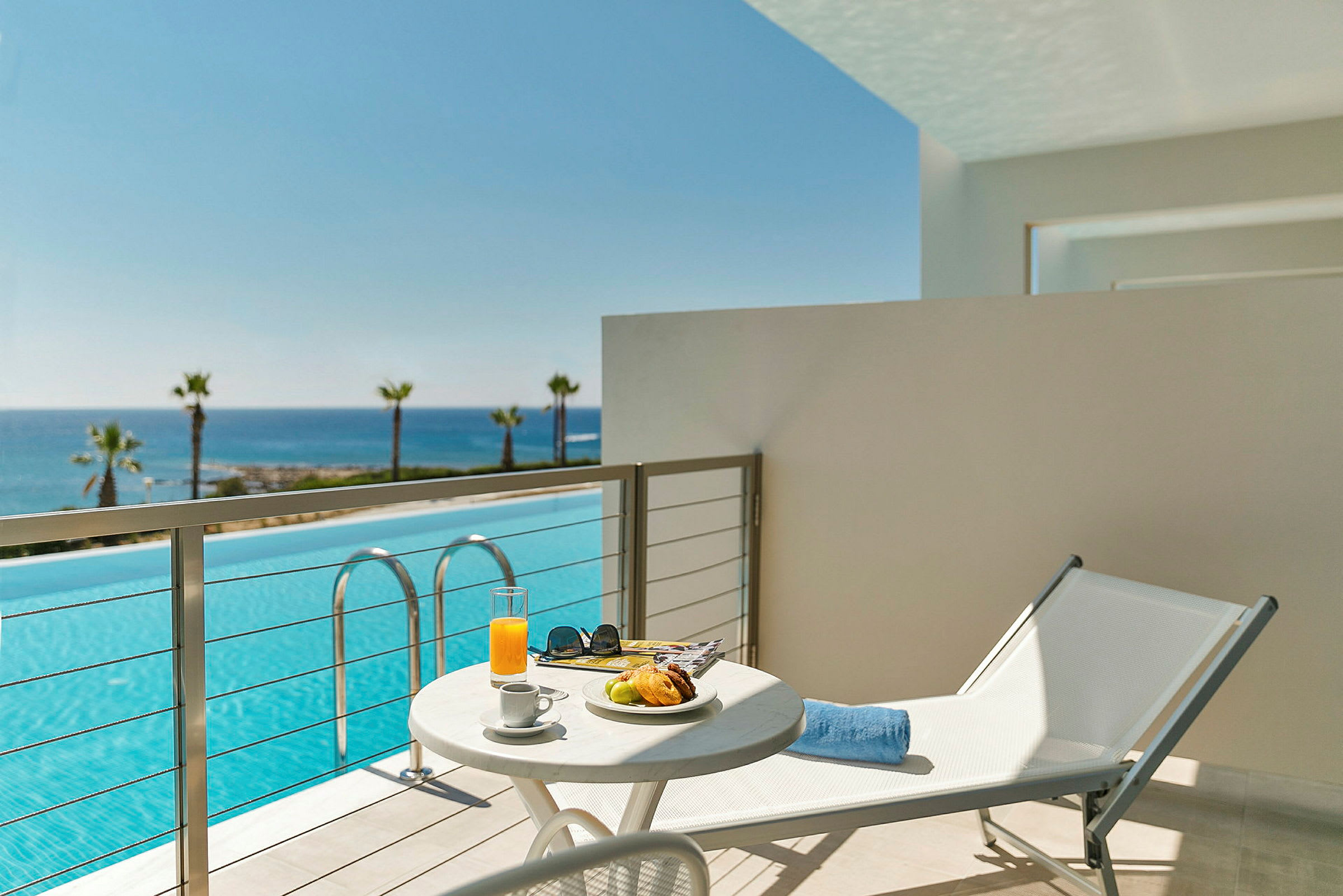 Balkong med utsikt över en pool och havet i bakgrunden, ett bord med frukost och en solstol. (AI-genererad text)