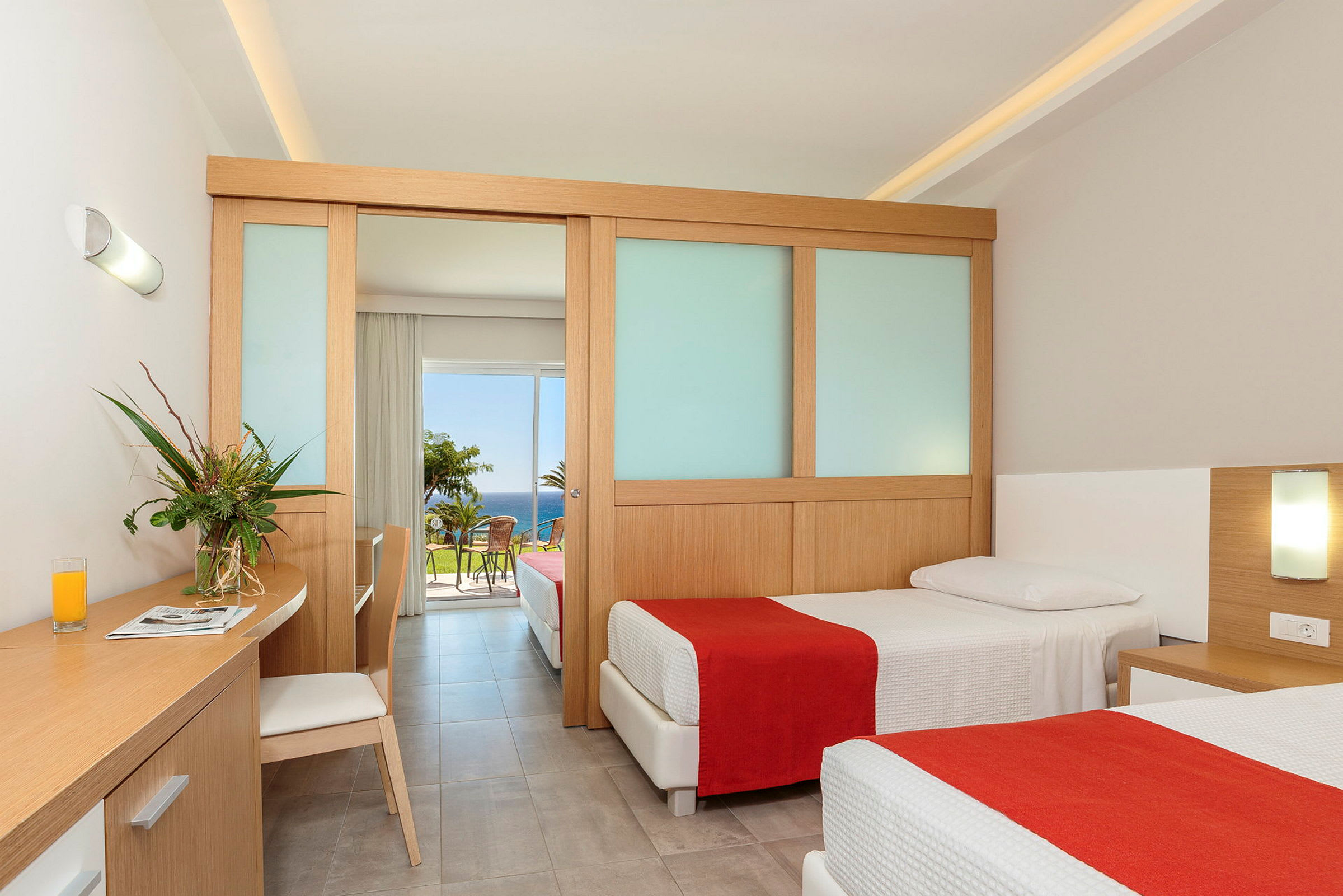 Ett hotellrum med två enkelsängar, en arbetsyta med en stol och en glasvas med blommor. Rummet har en skjutdörr som leder till en balkong med utsikt över havet. (AI-genererad text)