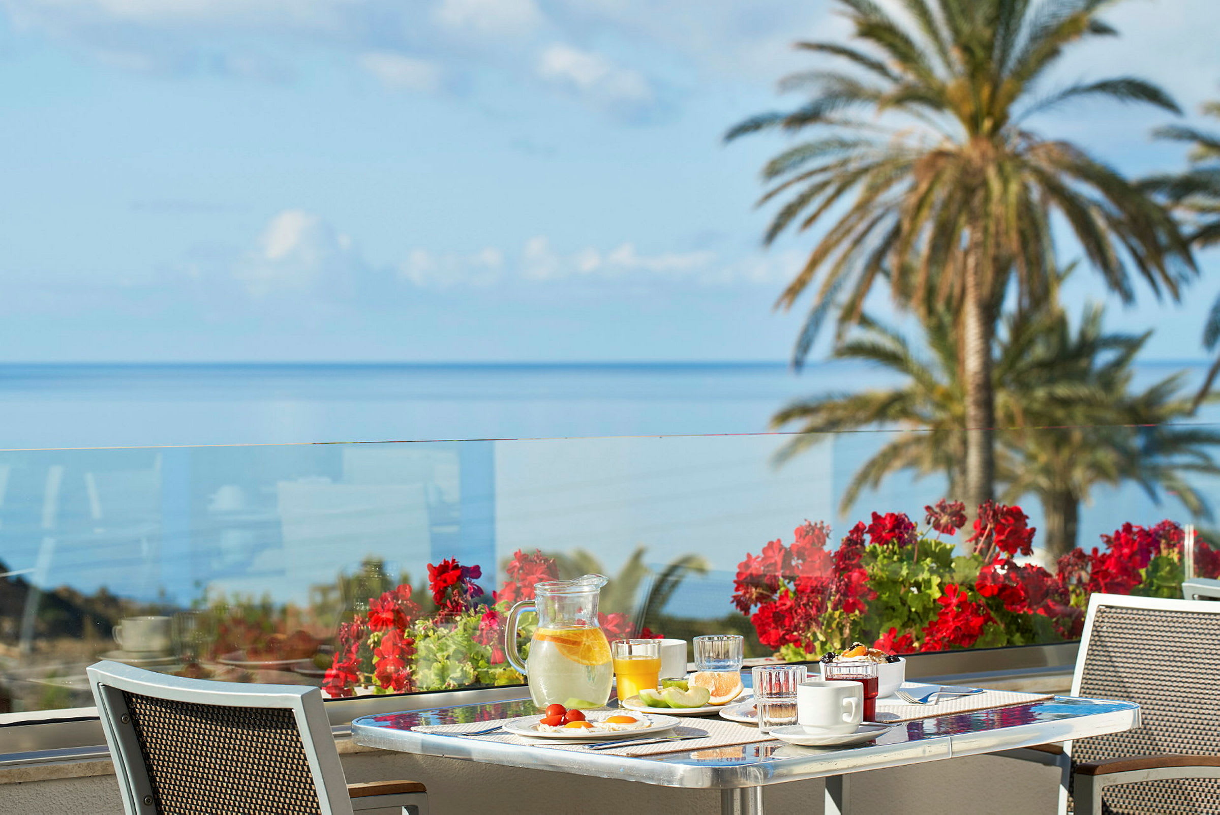En frukostservering på en terrass med havsutsikt och röda blommor i fokus. (AI-genererad text)