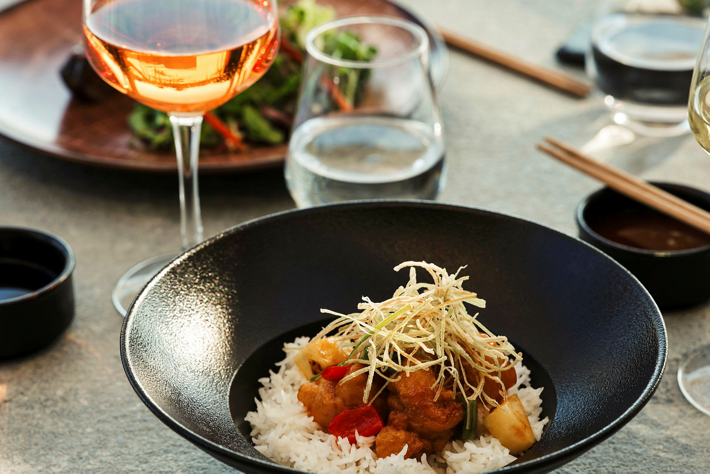 Närbild på en tallrik med asiatisk maträtt, serverad med ris och garnering, samt vinglas i bakgrunden. (AI-genererad text)