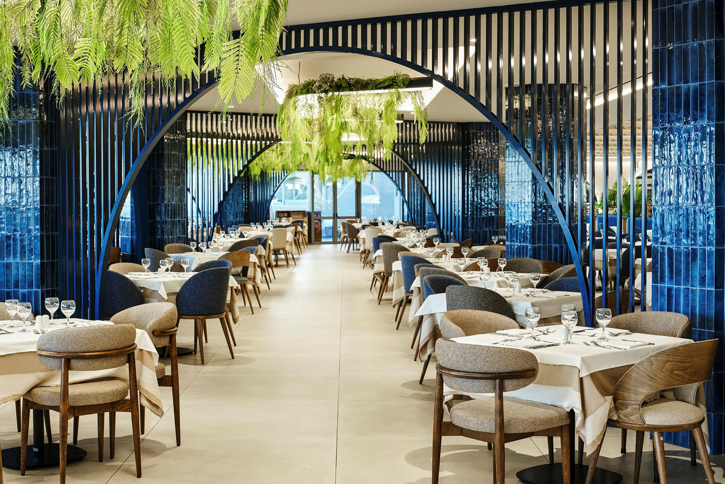 En elegant inredd restaurang med blåa bågstrukturer, vita dukar och gröna växter i taket. (AI-genererad text)