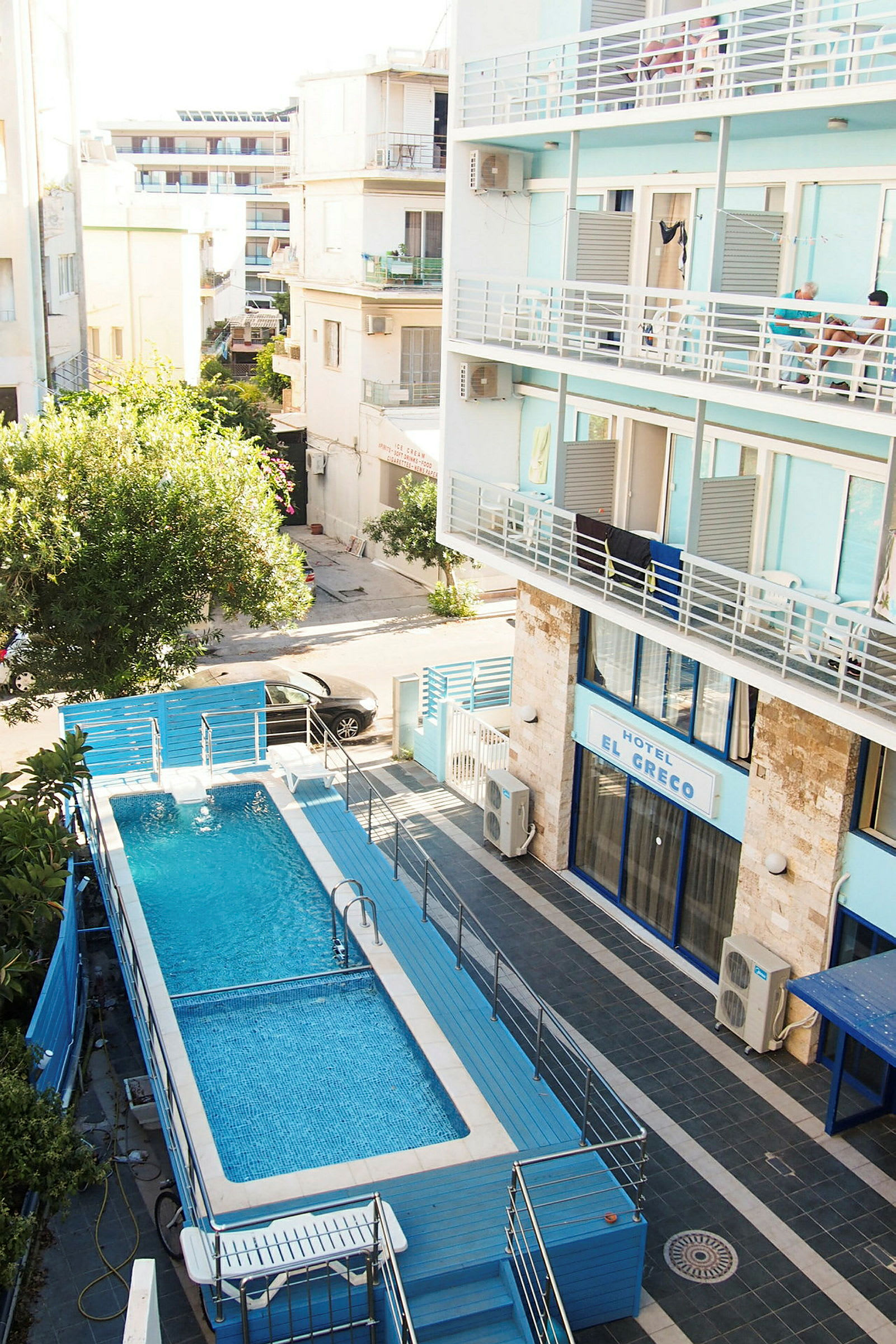 En swimmingpool med blå vatten omgiven av staket, belägen vid ett hotell med blå fasad. (AI-genererad text)