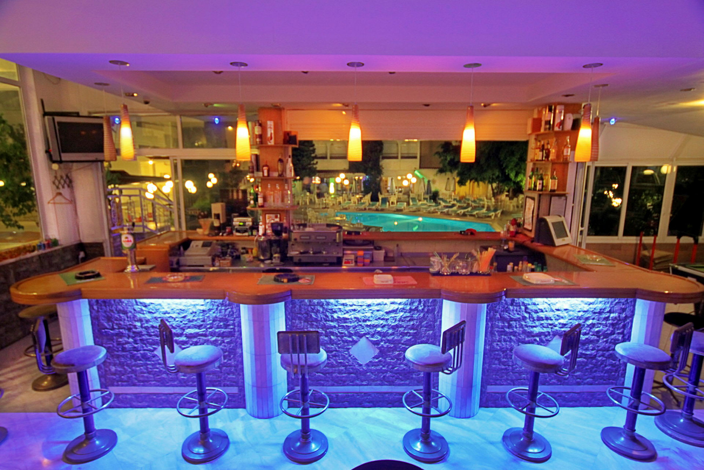 En upplyst bar med barstolar framför, och en bakgrund som visar en pool med solstolar. (AI-genererad text)