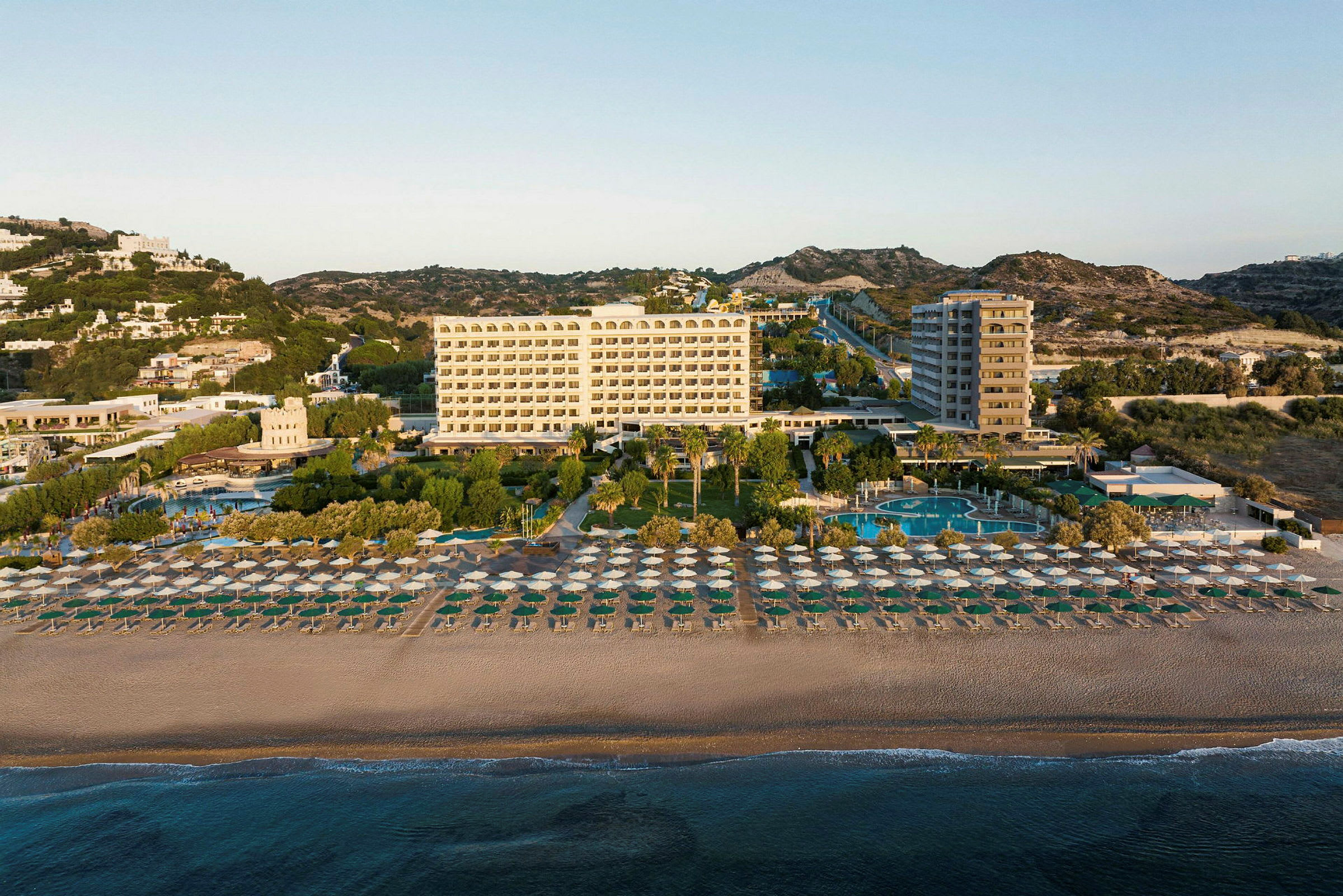 Ett hotell med flera våningar bakom en sandstrand fylld med solparasoller, omgiven av träd och pooler. (AI-genererad text)