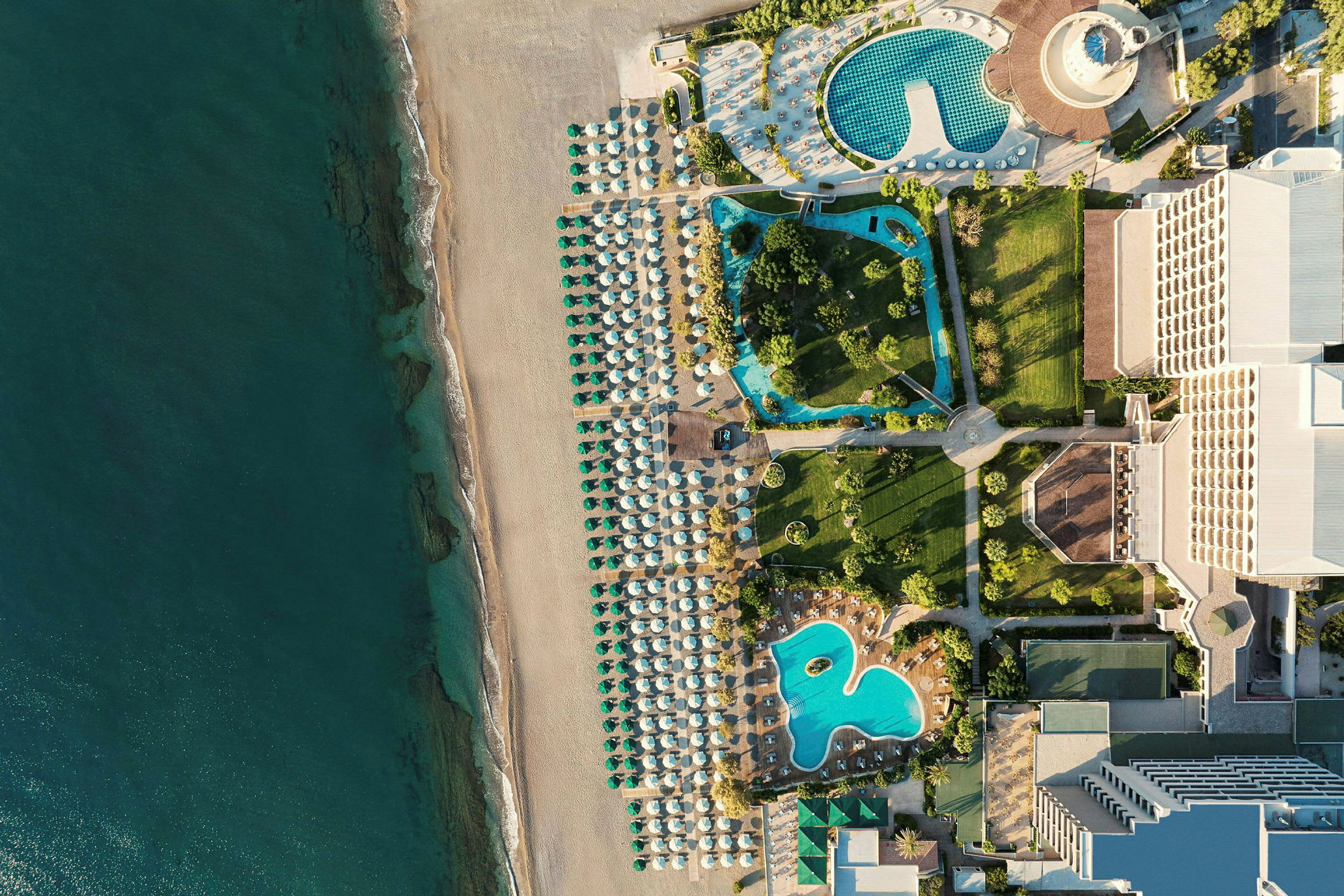 Flygfoto som visar en strand med parasoller, poolområden och hotellbyggnader. (AI-genererad text)