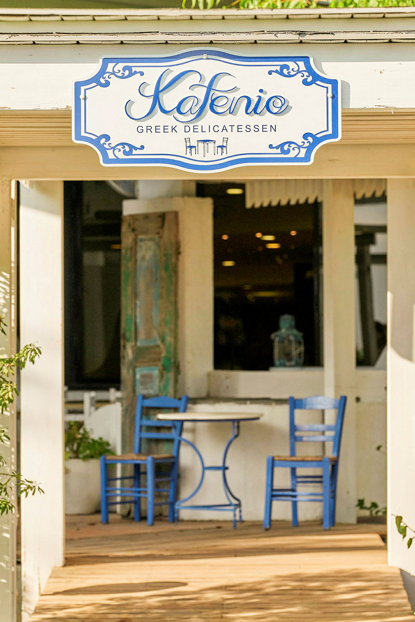 Två blå stolar och ett runt bord står på en utomhusterrass framför en byggnad. En skylt ovanför dörren lyder "Kafenio Greek Delicatessen". (AI-genererad text)