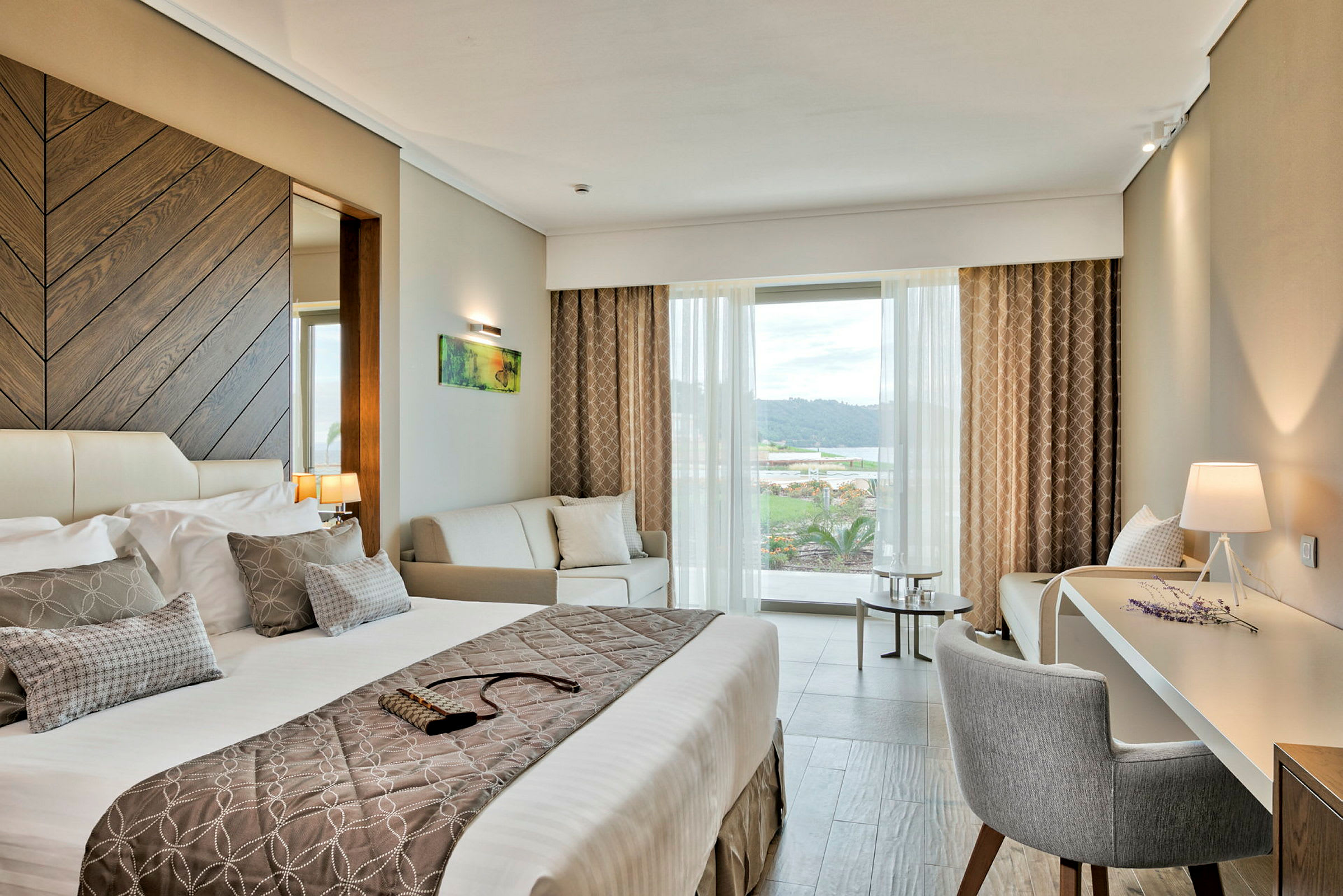 Ett modernt hotellrum med en stor säng, en soffa och ett skrivbord med en lampa bredvid. Fönstret visar en utsikt över ett landskap. (AI-genererad text)