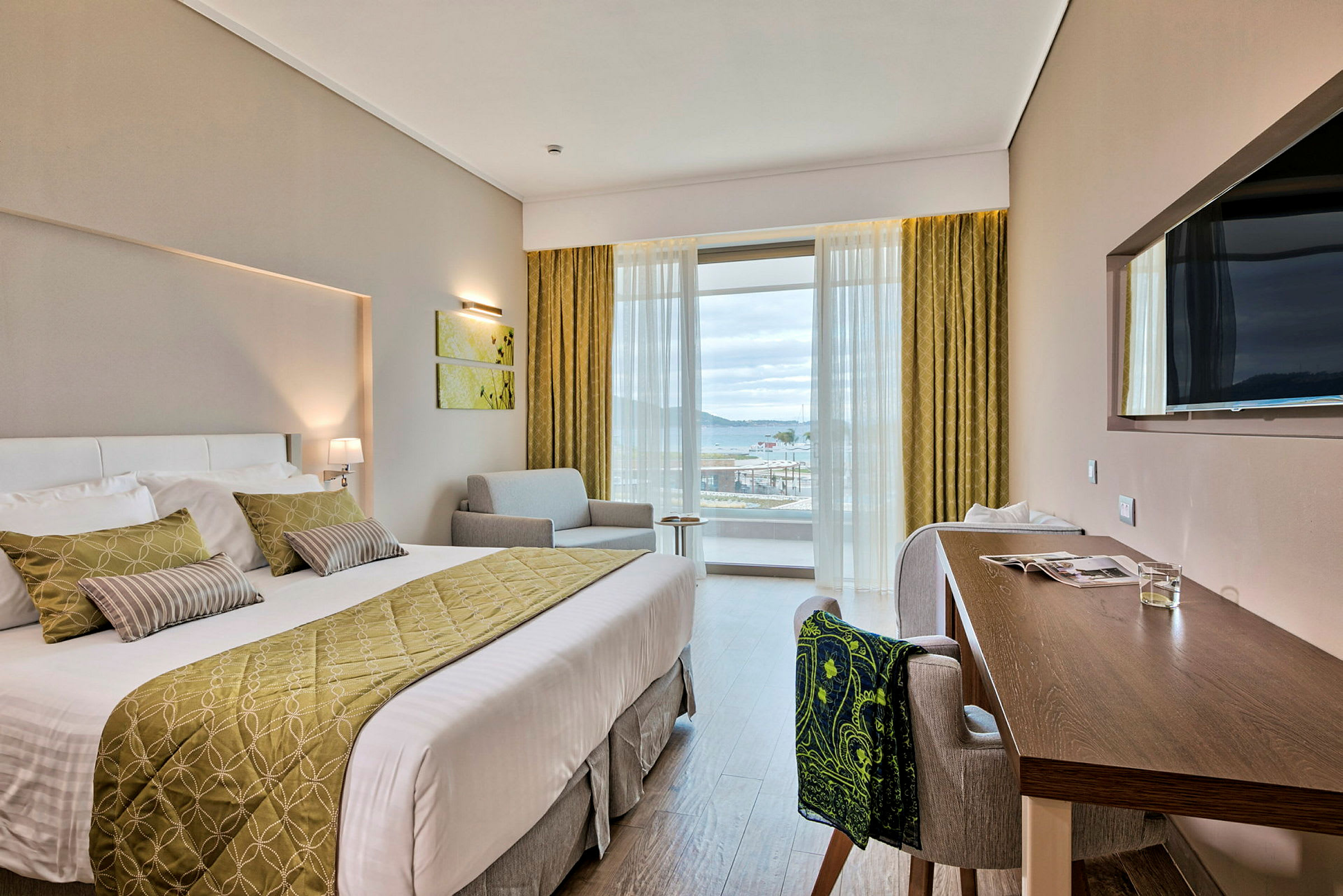 Ett hotellrum med en dubbelsäng, ett skrivbord och en sittmöbel. Ett stort fönster ger utsikt mot en hamn. (AI-genererad text)