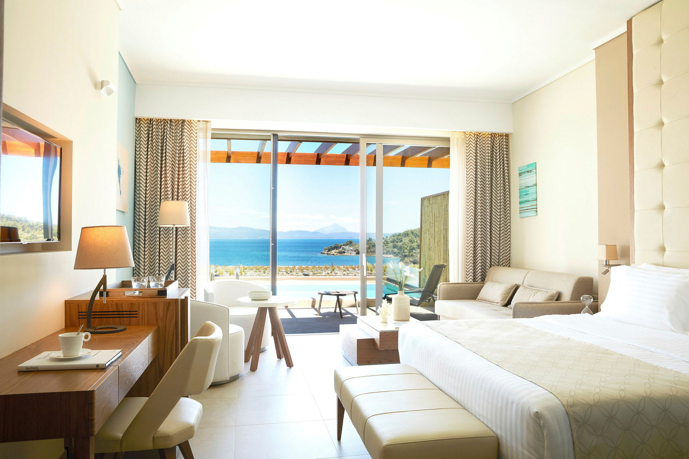 Ett hotellrum med modern inredning och stora glasdörrar som öppnar upp mot en terrass med utsikt över havet. (AI-genererad text)
