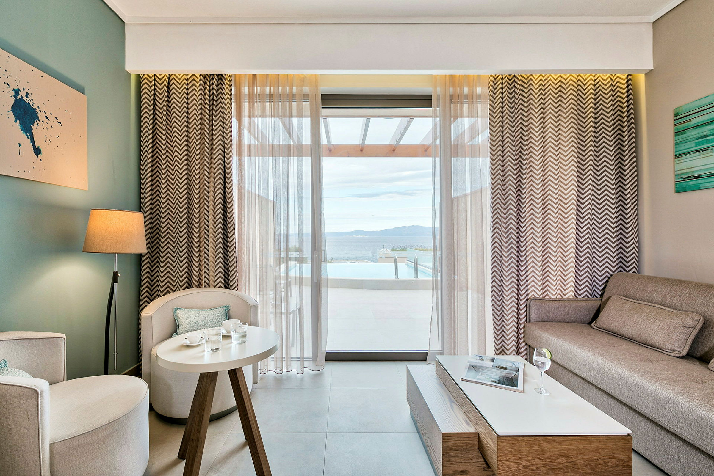 Bilden visar ett vardagsrum med modern inredning, en soffa, två fåtöljer, ett litet bord och ett stort fönster som vetter mot en veranda och utsikt över havet. (AI-genererad text)