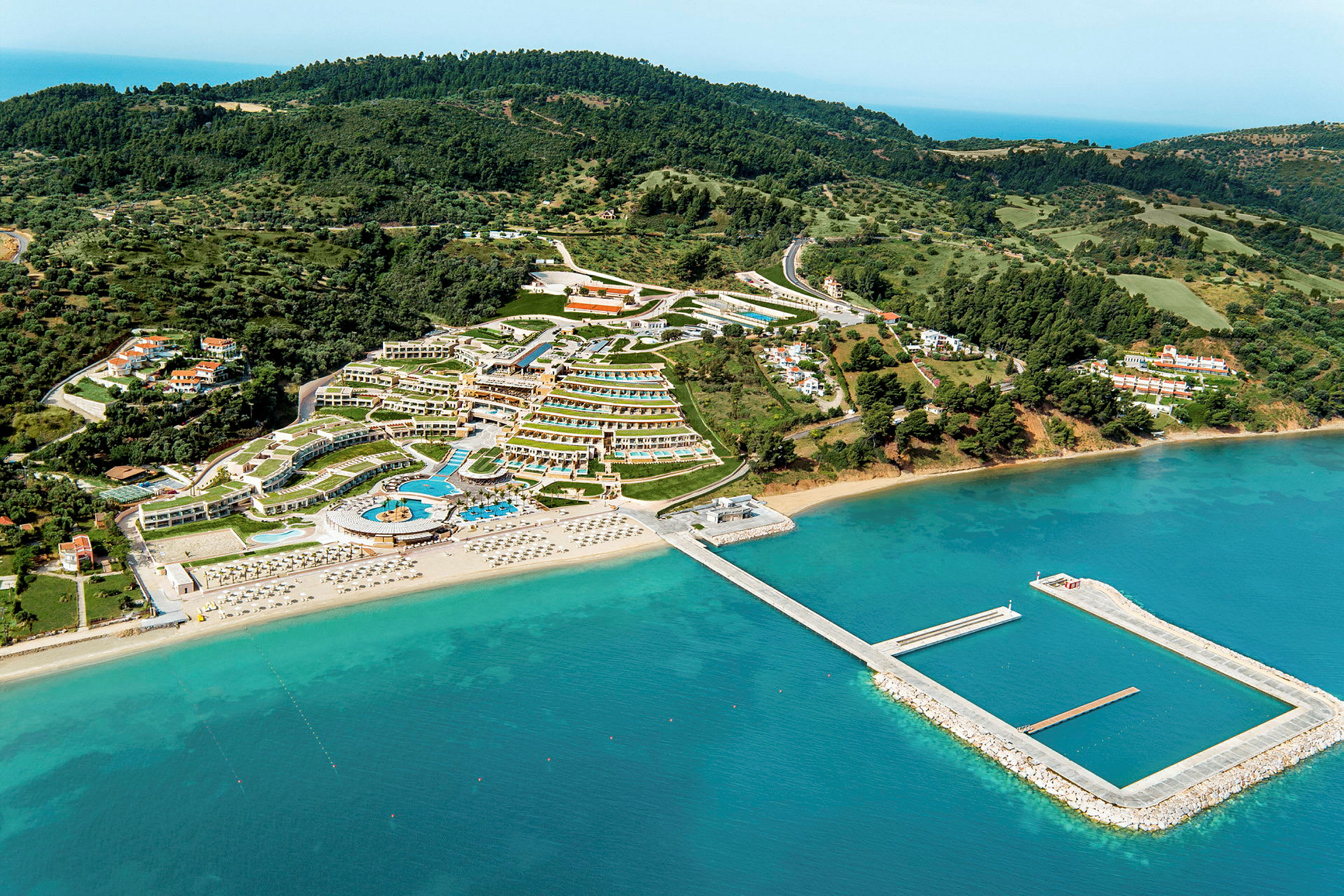 Flygbild över ett semesterområde med hotell, pooler och strand med solstolar, omgivet av grönska och hav. (AI-genererad text)