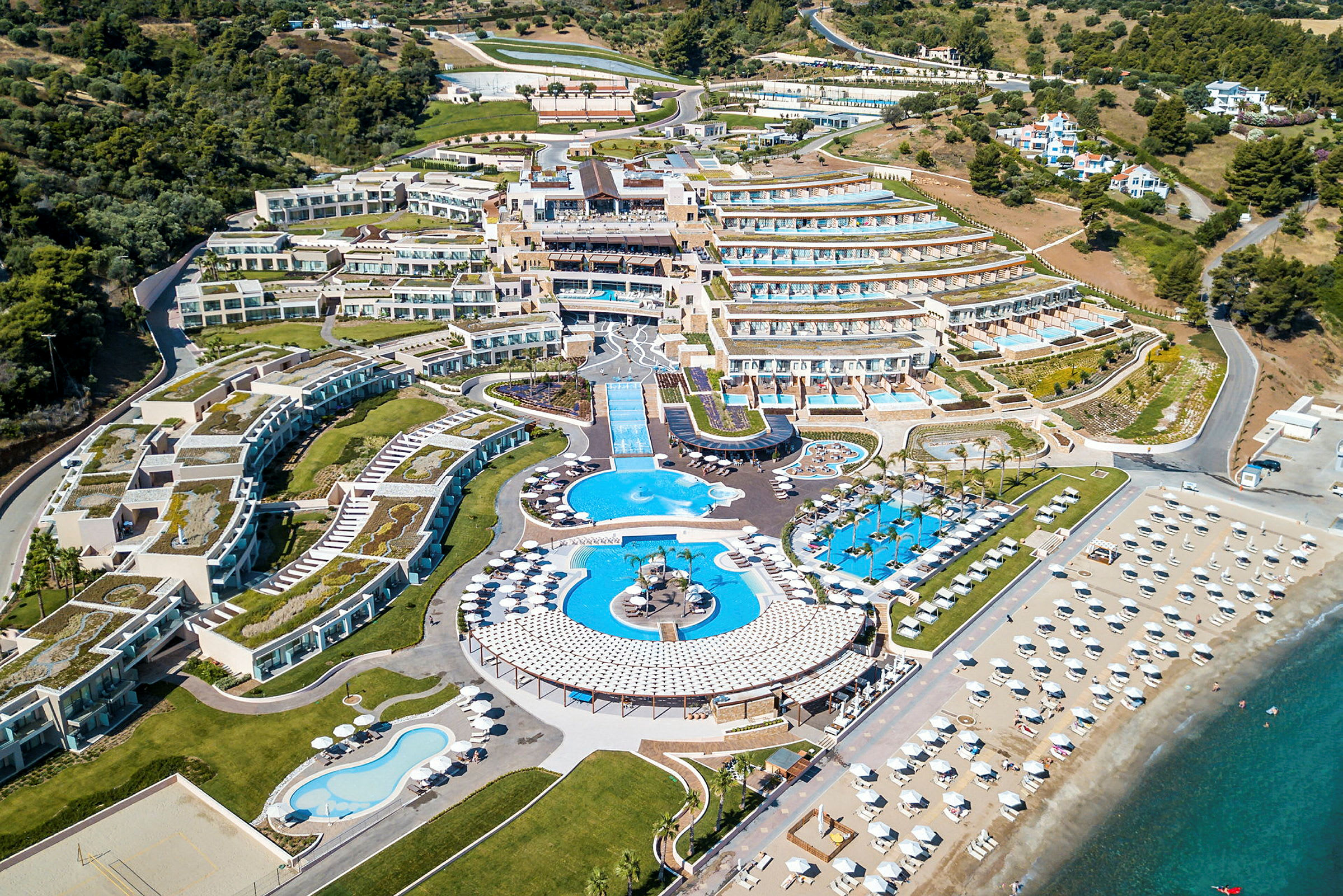 Flygbild över ett stort resortområde med terrasserade byggnader, flera pooler och en bred sandstrand med parasoller. (AI-genererad text)