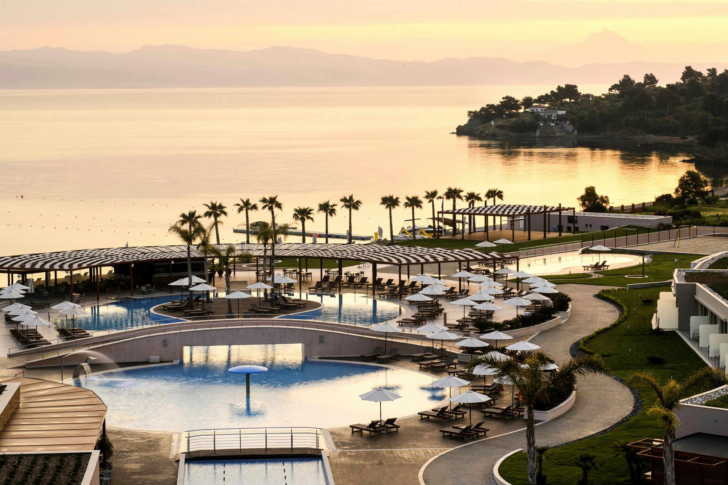 En resort med poolområde, solstolar och parasoller vid kusten, med havsutsikt och solnedgång. (AI-genererad text)