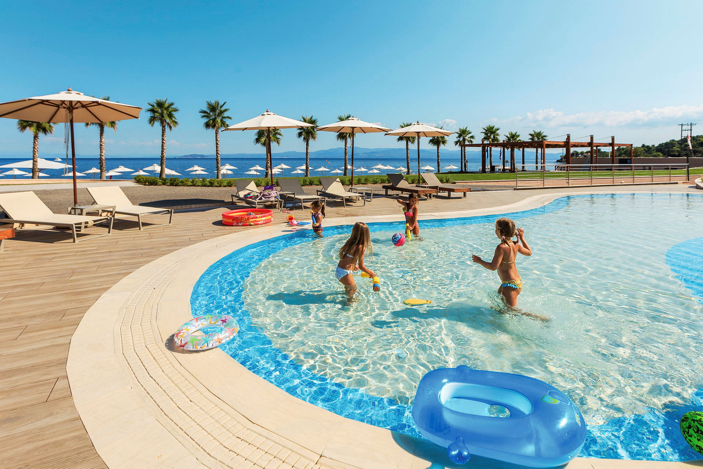 Barn leker i en grund pool vid en resort nära havet. Solstolar och parasoller finns runt poolområdet. (AI-genererad text)