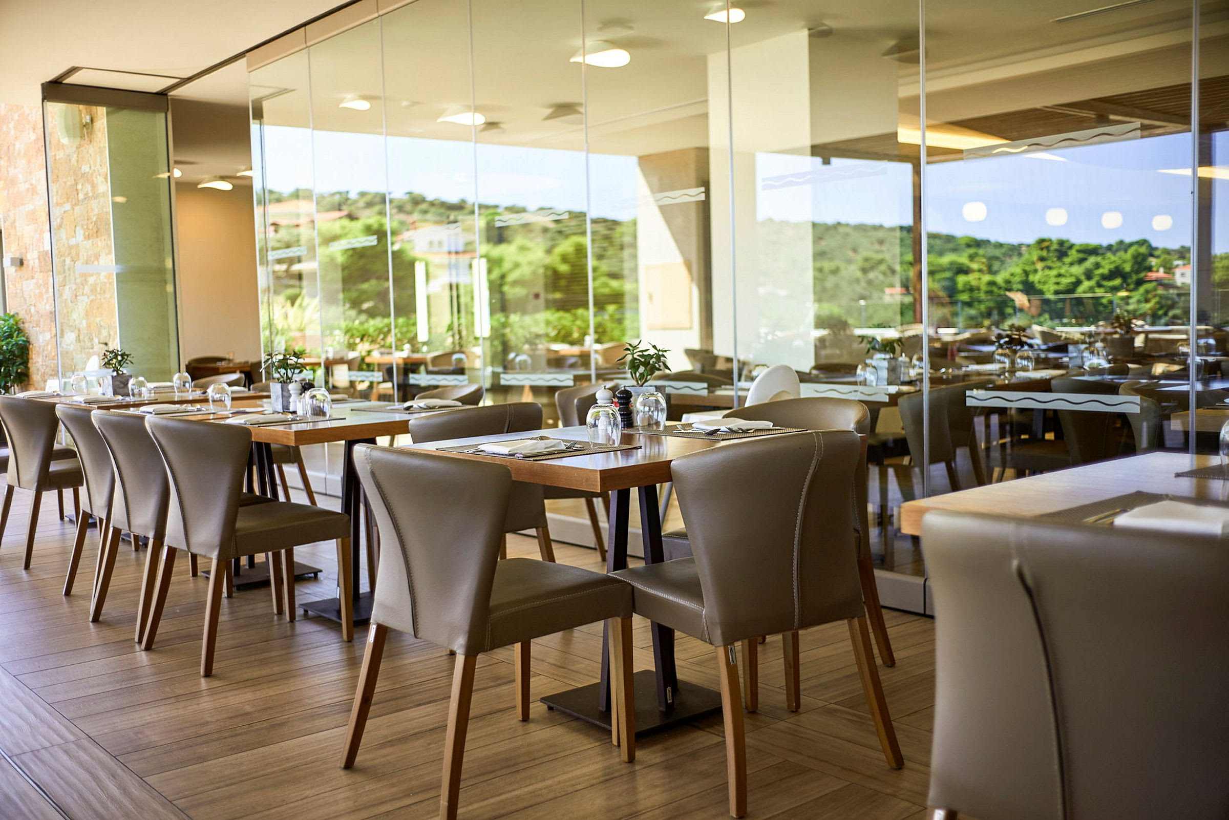 Ett modernt restaurangområde med bord och stolar samt utsikt genom glasväggarna mot grönska. (AI-genererad text)