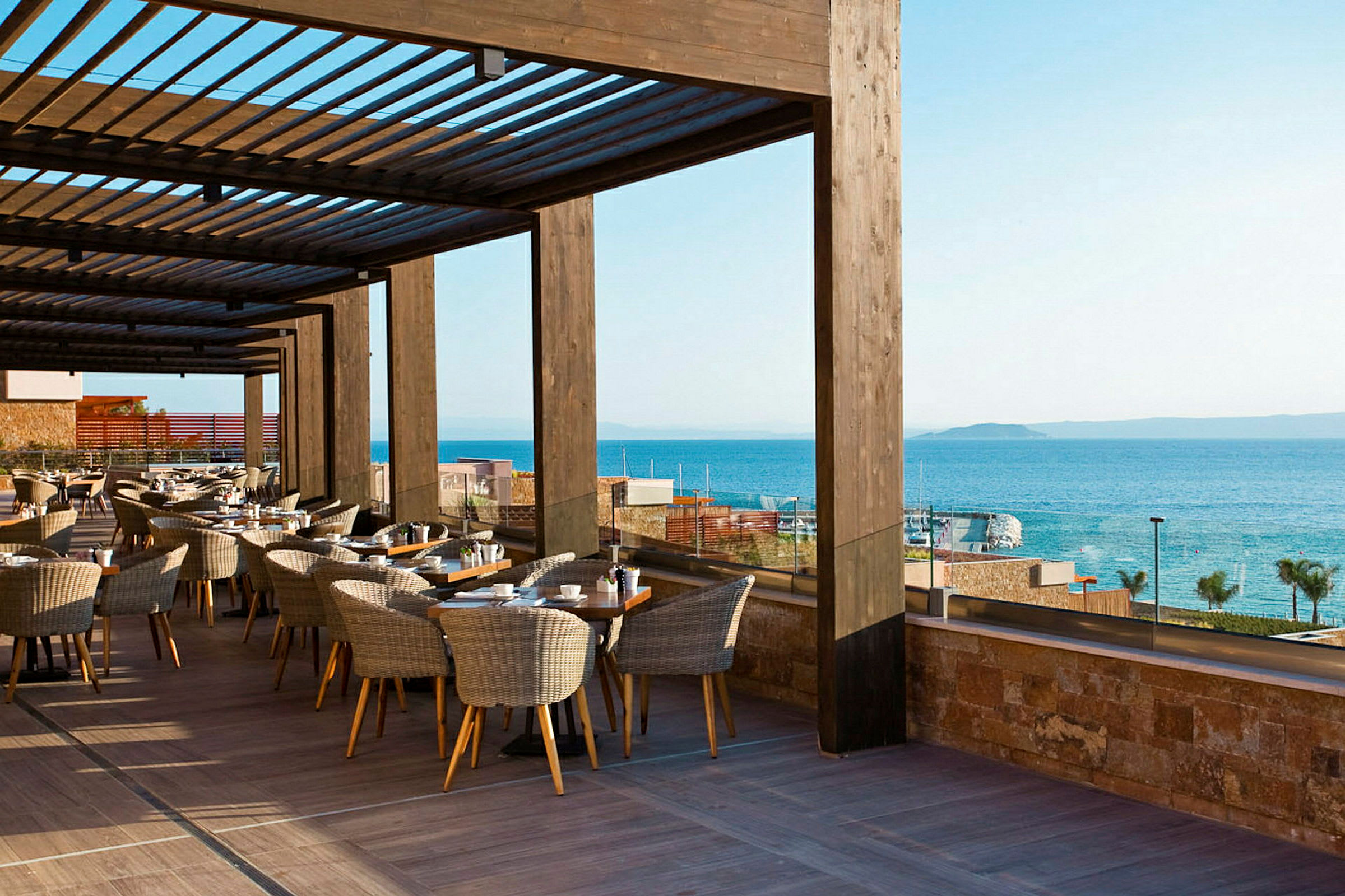 En terrass med bord och stolar under en träpergola med utsikt över havet i bakgrunden. (AI-genererad text)