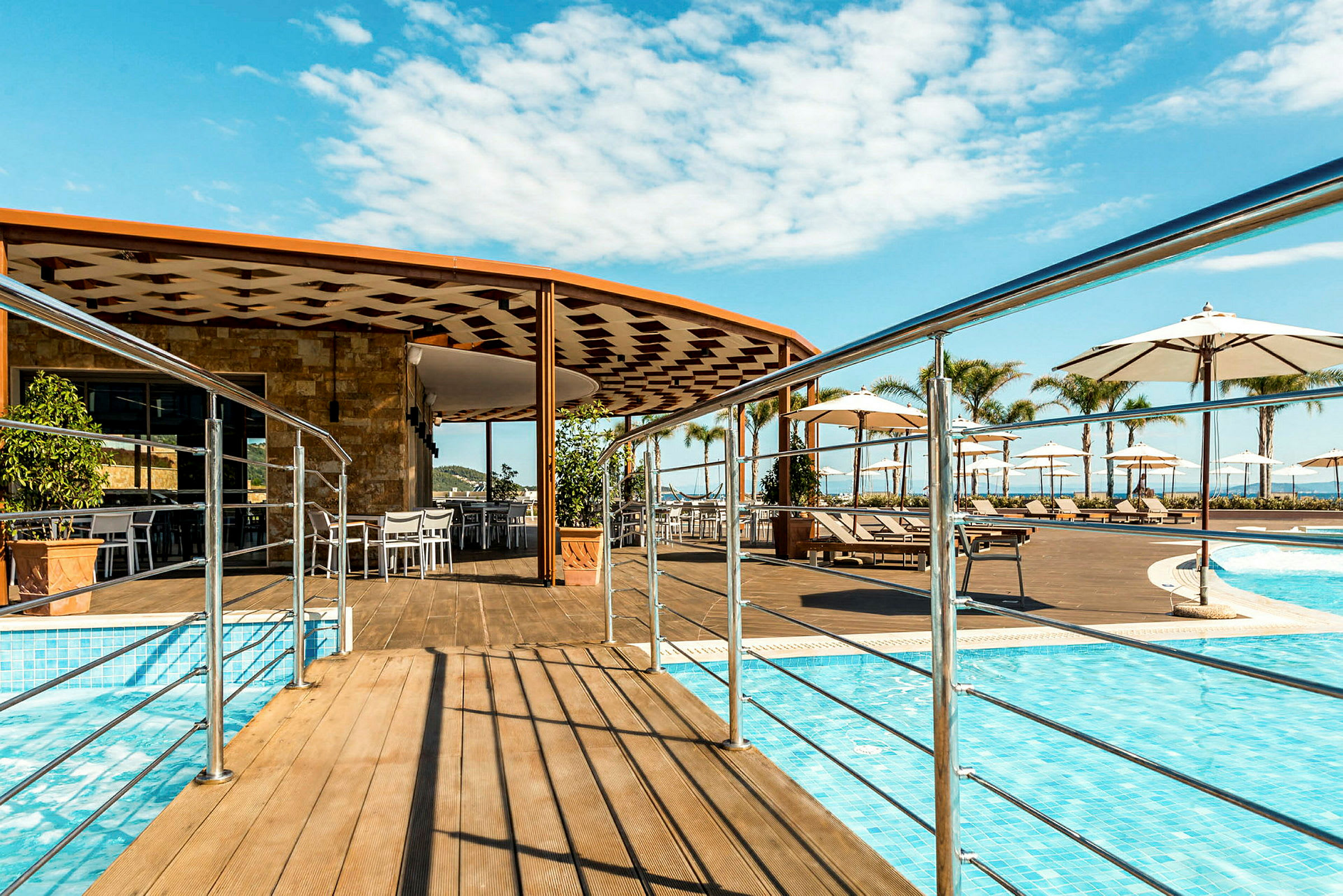 En utomhuspool med en träterrass, parasoller och solsängar. I bakgrunden syns en byggnad med uteservering. (AI-genererad text)