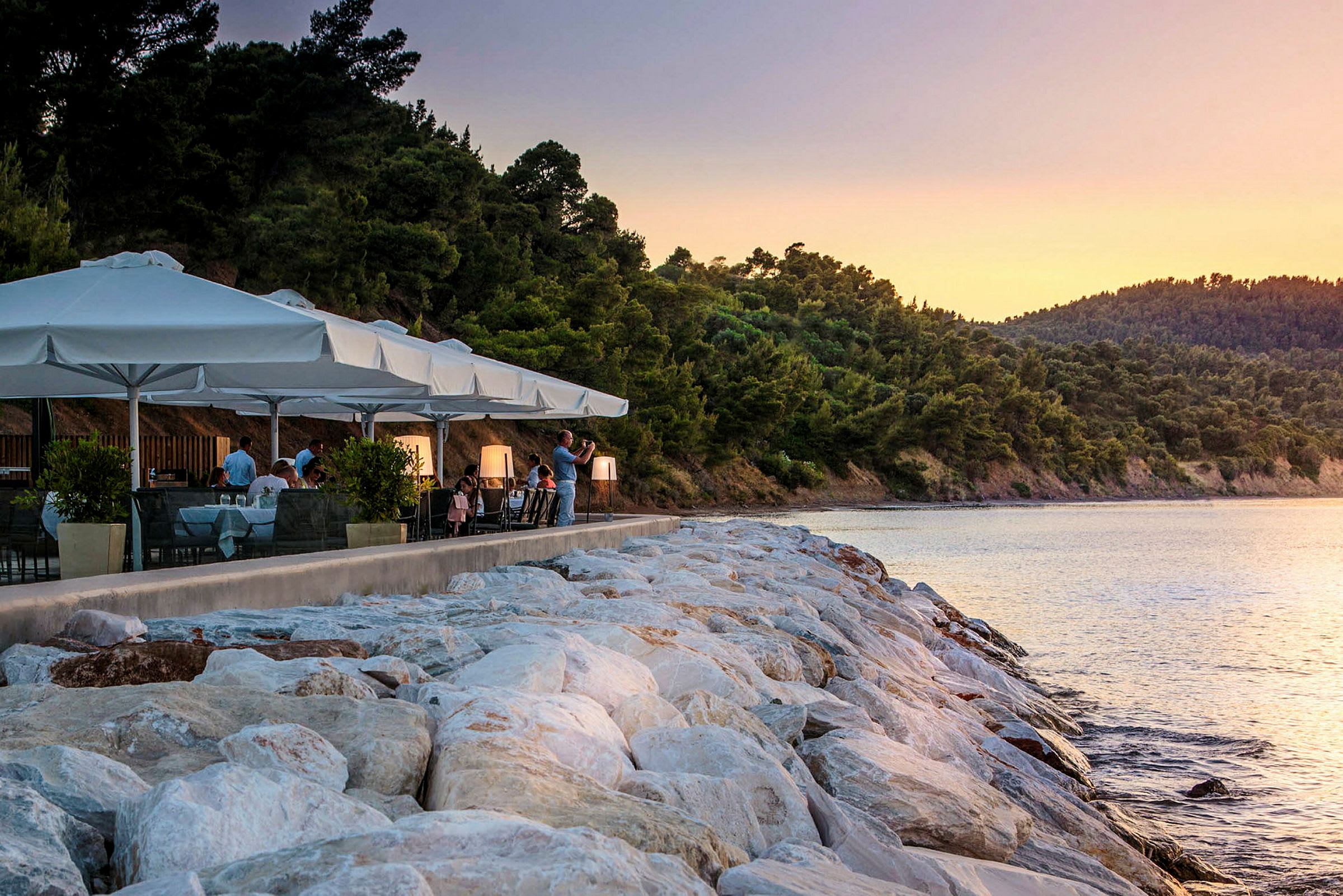 En restaurang med vita parasoller vid stranden, omgiven av grönska och vatten med solnedgång. (AI-genererad text)