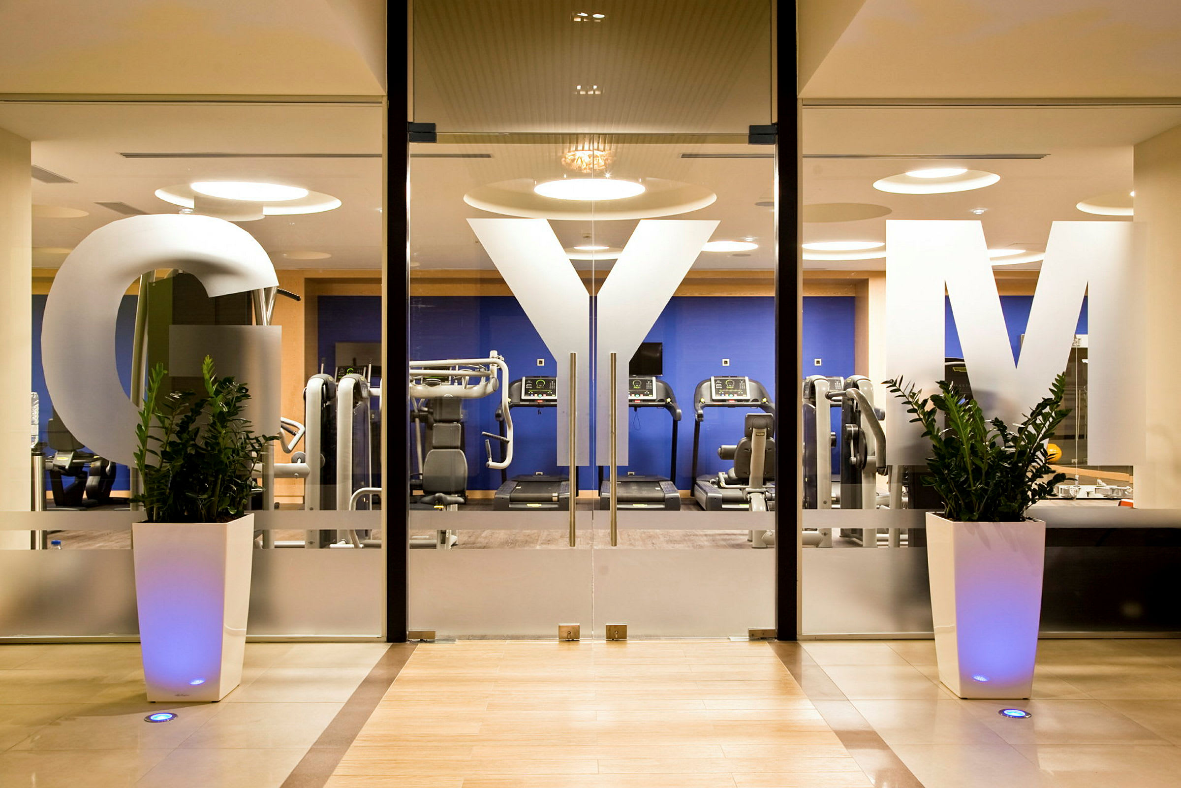 En entré till ett gym med glasdörrar prydda med bokstäverna "GYM" och synliga maskiner och träningsutrustning i bakgrunden. (AI-genererad text)