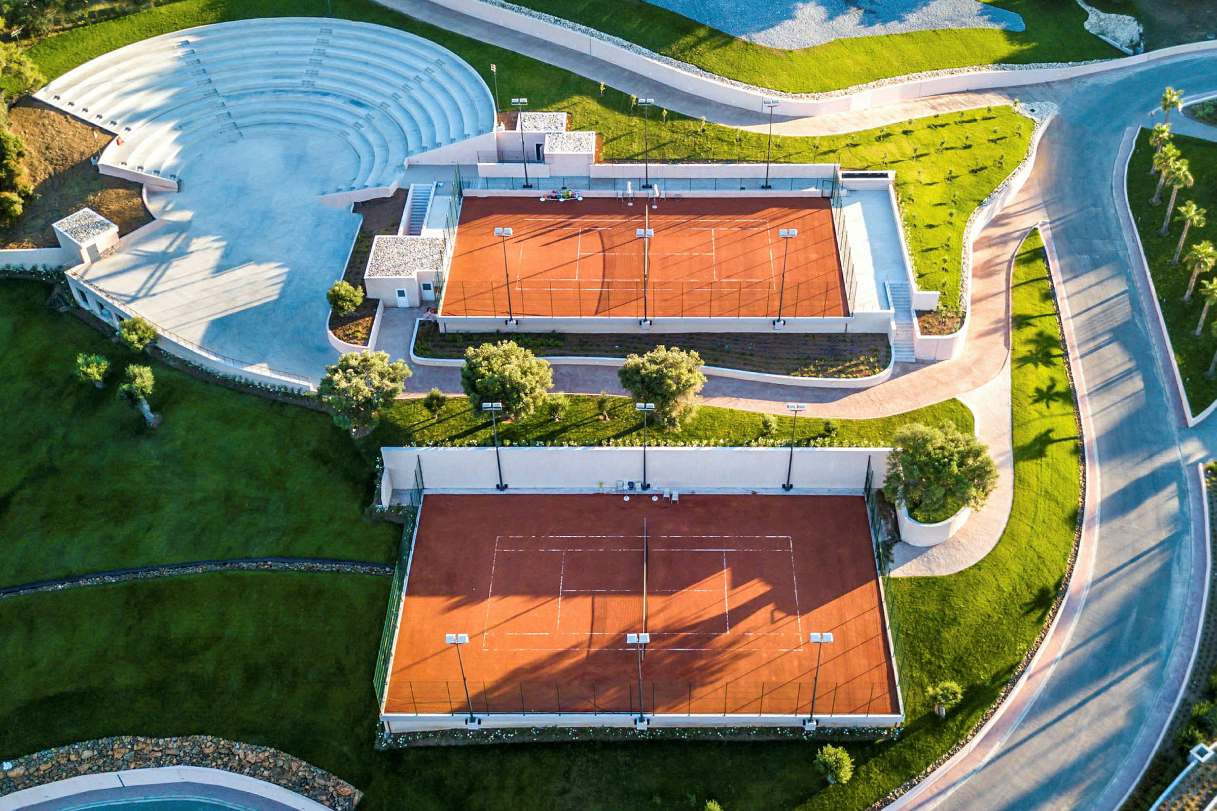 Flygbild över två tennisbanor med rött underlag och ett angränsande utomhusamfiteater. (AI-genererad text)