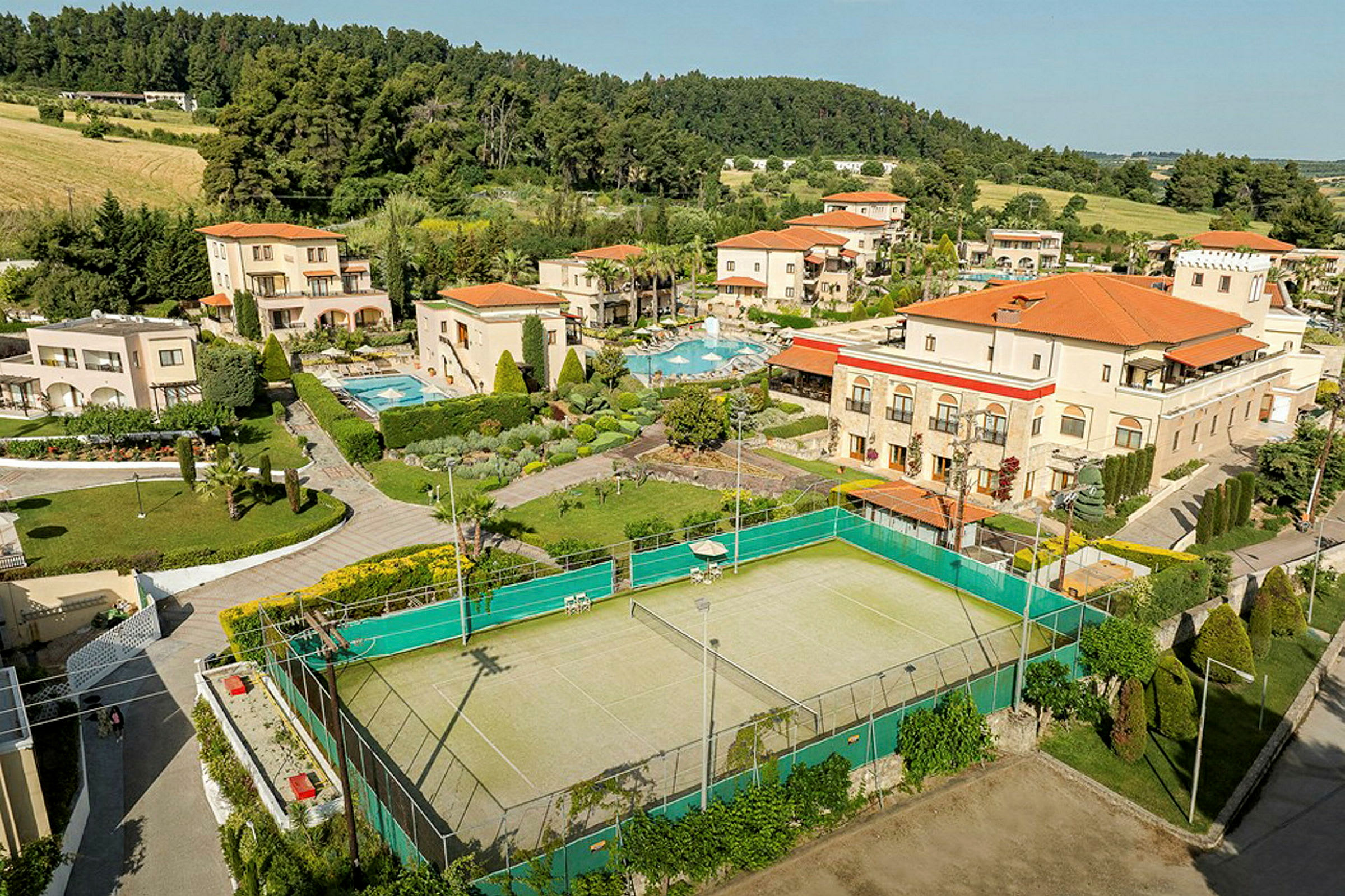 Flygbild av ett semesterkomplex med pooler, trädgårdar och en tennisbana, omgivet av grönska och skog. (AI-genererad text)