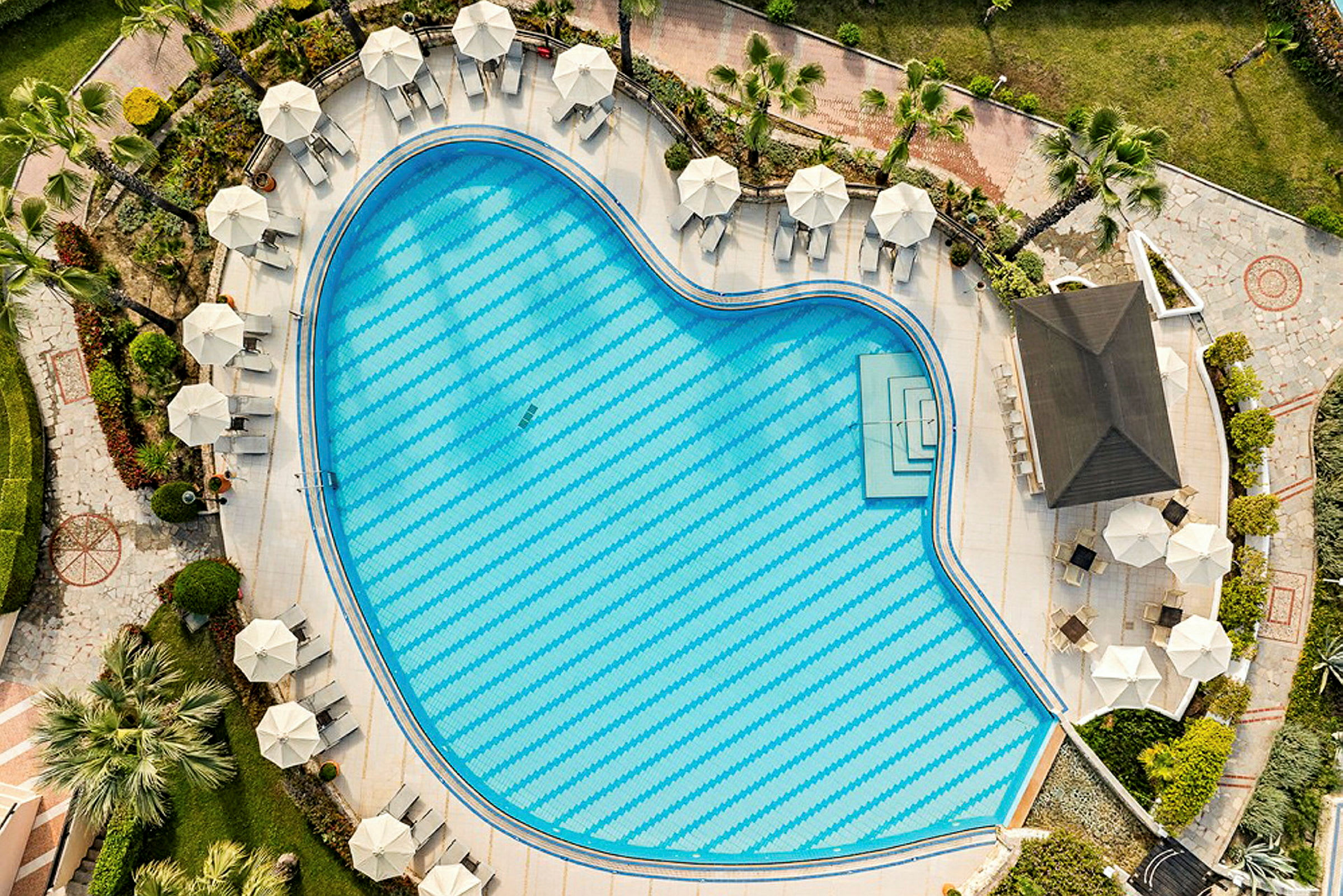 Flygfoto av en hjärtformad pool omgiven av parasoller och solstolar placerade på ett välskött poolområde. (AI-genererad text)