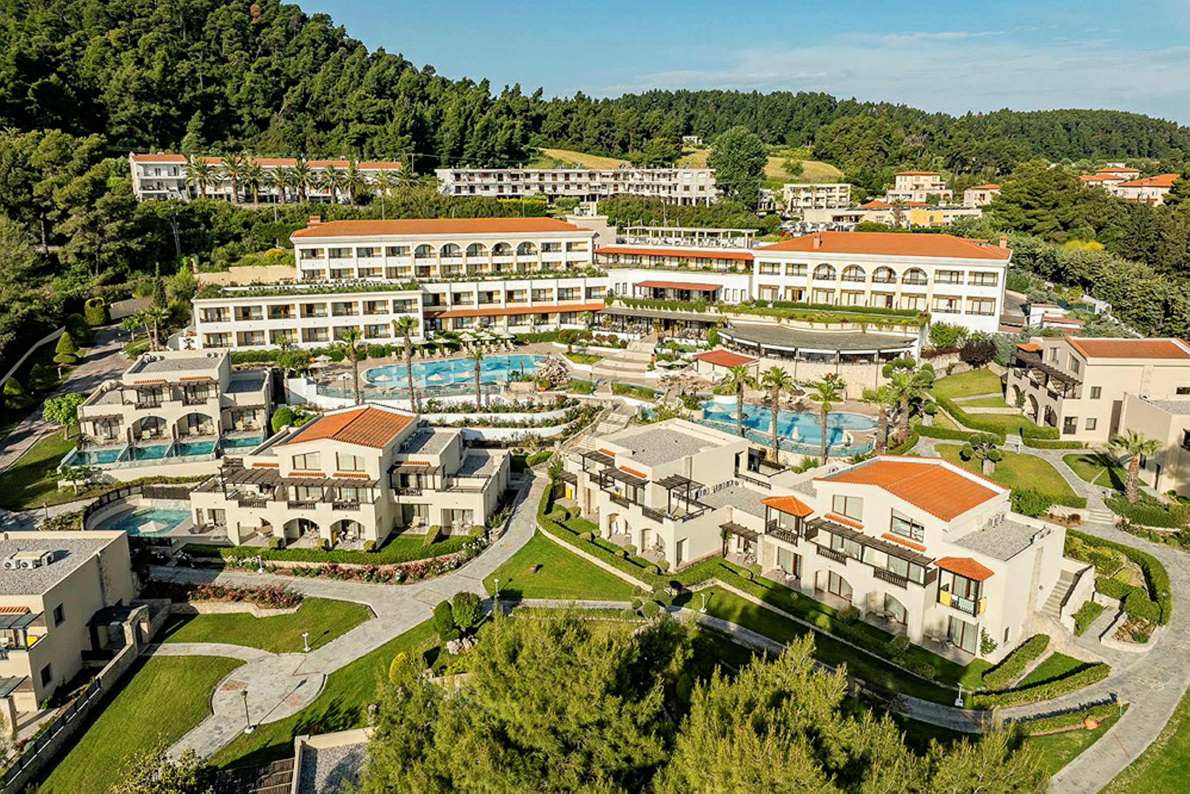 En bild av ett semesterresort som består av flera byggnader med vita fasader och röda tak. Resorten har poolområden och är omgiven av grönska och träd. (AI-genererad text)