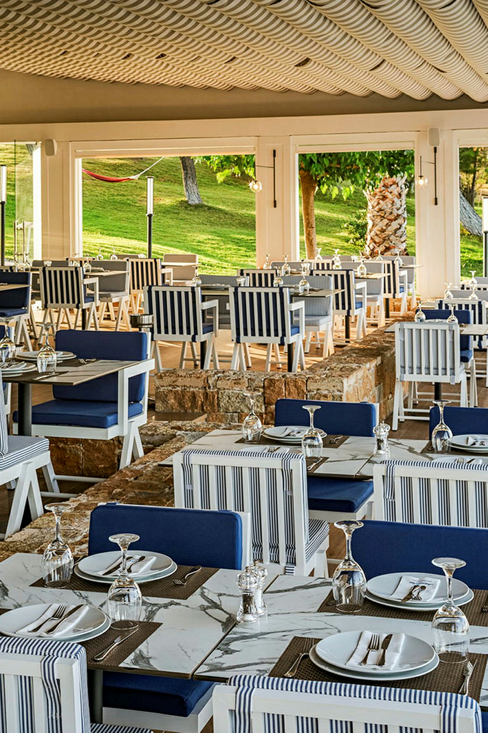 En restaurang med uppdukade bord och stolar, dekorerade i blå och vita färger, med en utsikt över en gröngräsbevuxen utomhusmiljö. (AI-genererad text)